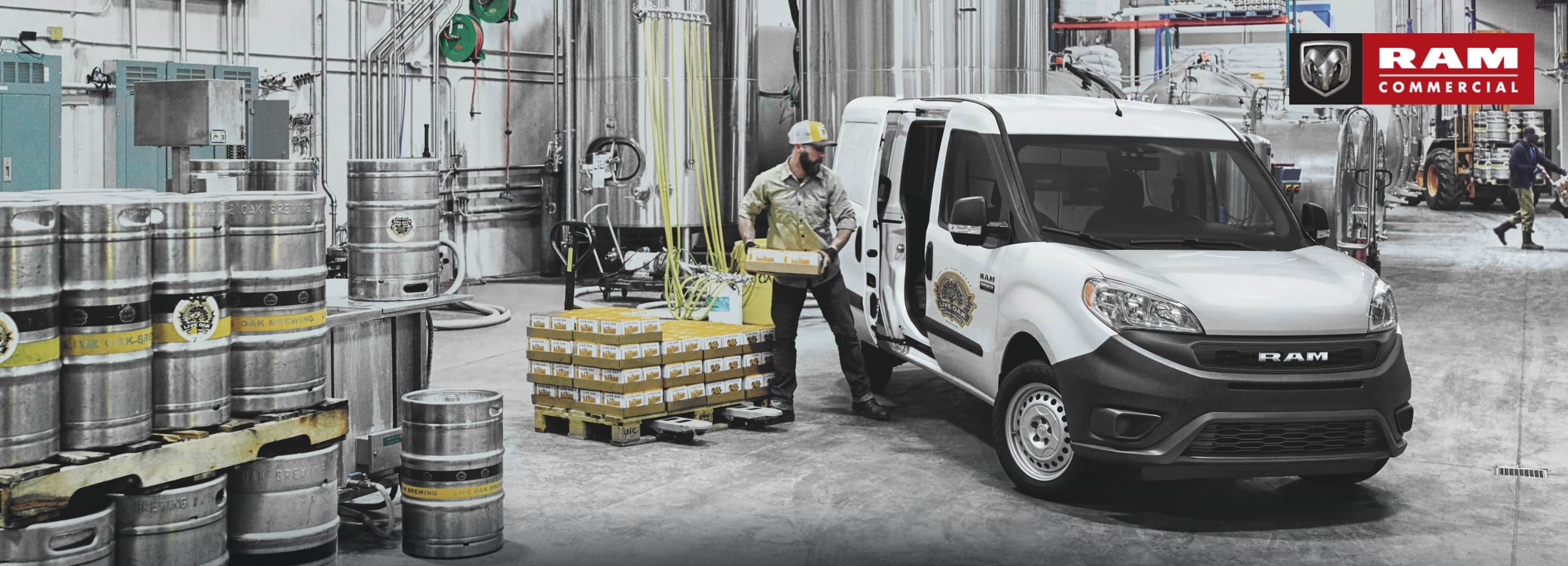 Una Cargo Van Ram ProMaster City Tradesman 2021 estacionada en un almacén y un hombre cargando cajas de artículos a través de la puerta deslizable lateral. El logo de la Ram comercial.