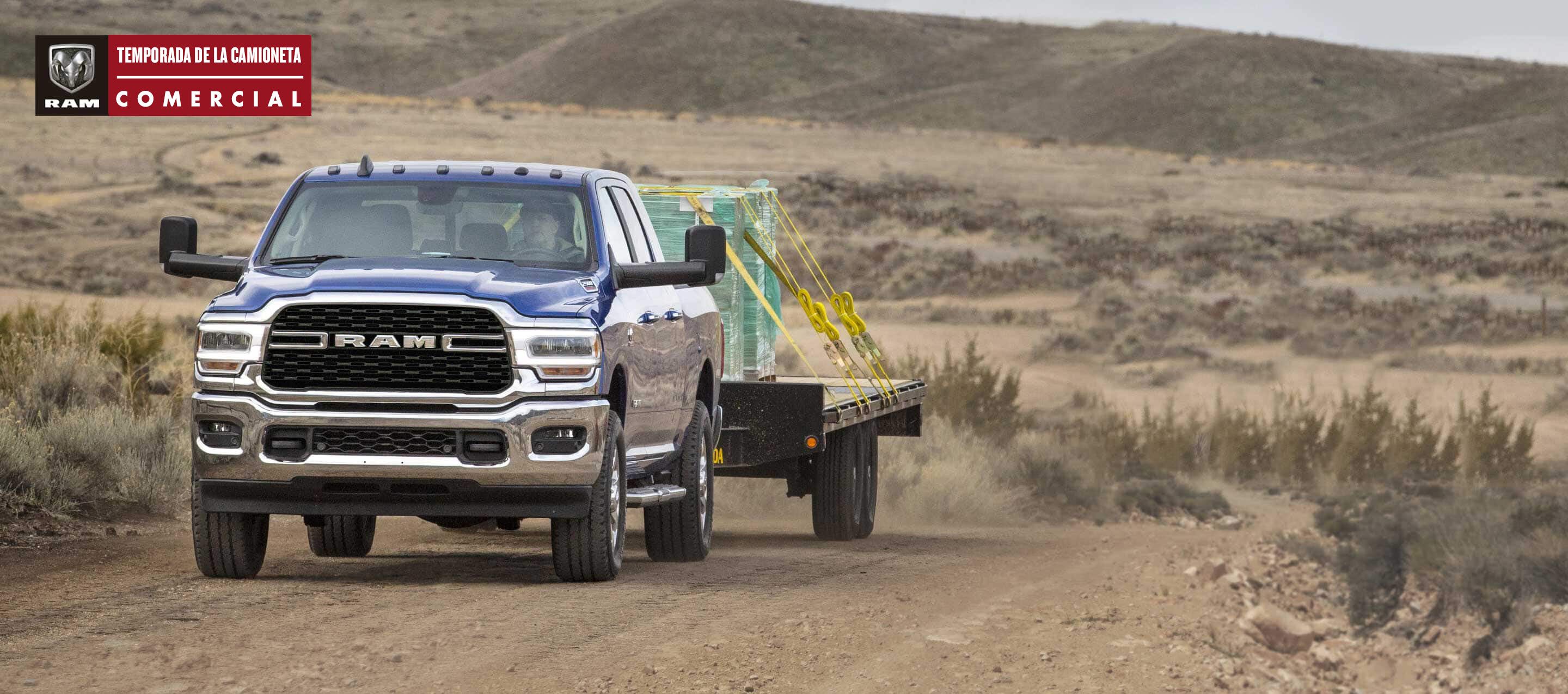 Una Ram 2500 Big Horn 4x4 Crew Cab 2022 azul circulando por una carretera sinuosa en el desierto, acarreando paneles de vidrio en un remolque de plataforma plana. Temporada de la camioneta comercial Ram.
