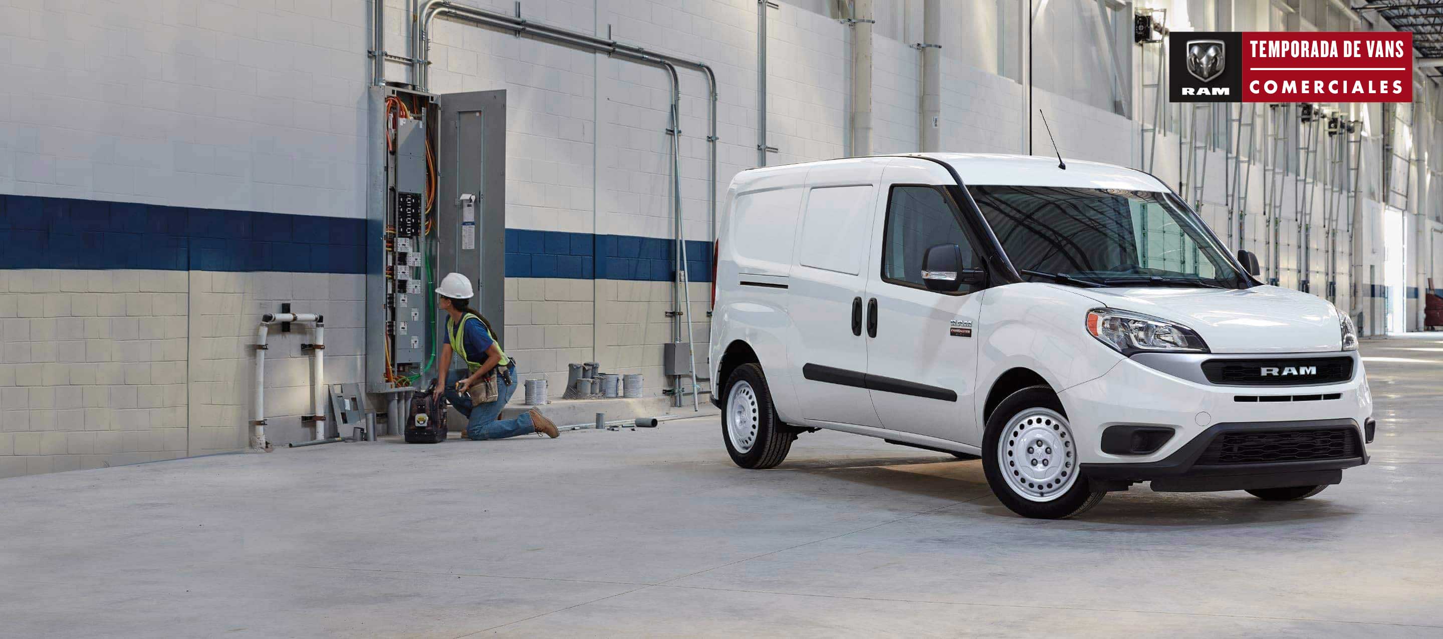 Una Ram ProMaster City Cargo Van 2022 blanca estacionada en un gran garaje industrial, con un técnico cerca que trabaja en una caja de disyuntores eléctricos. Publicidad de Ram.