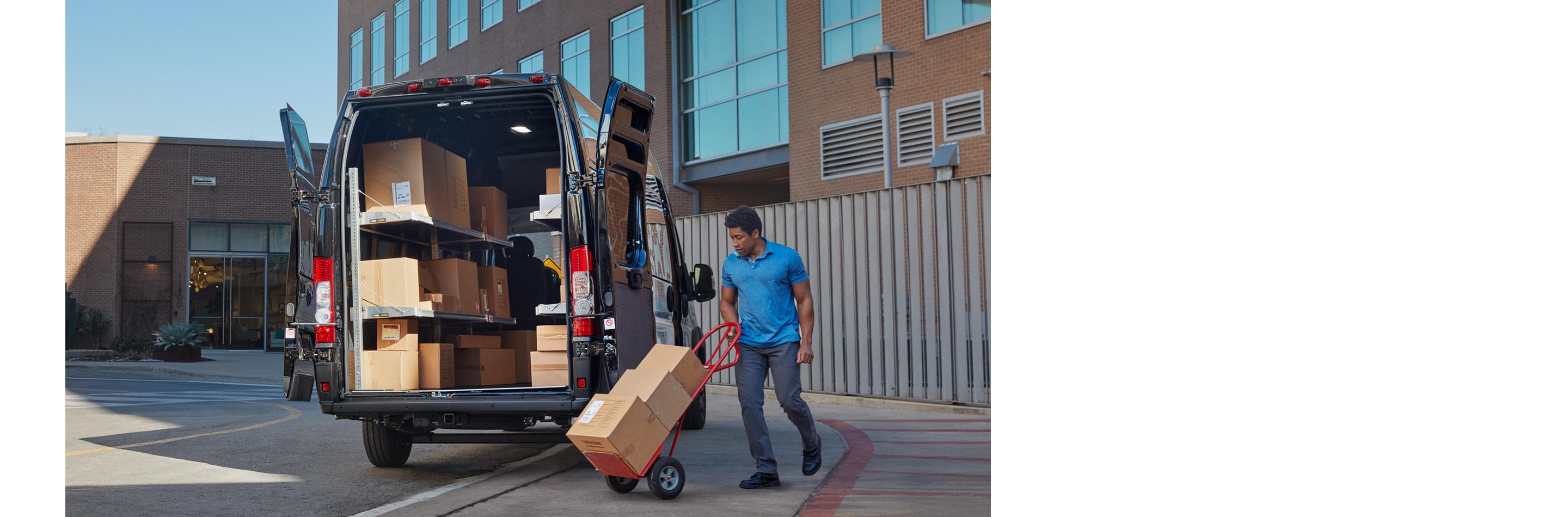 Hombre llevando paquetes en una carretilla en dirección hacia las puertas traseras abiertas de una Ram ProMaster 3500 SLT Cargo Van Super High Roof 2024 con el área de carga llena de cajas.