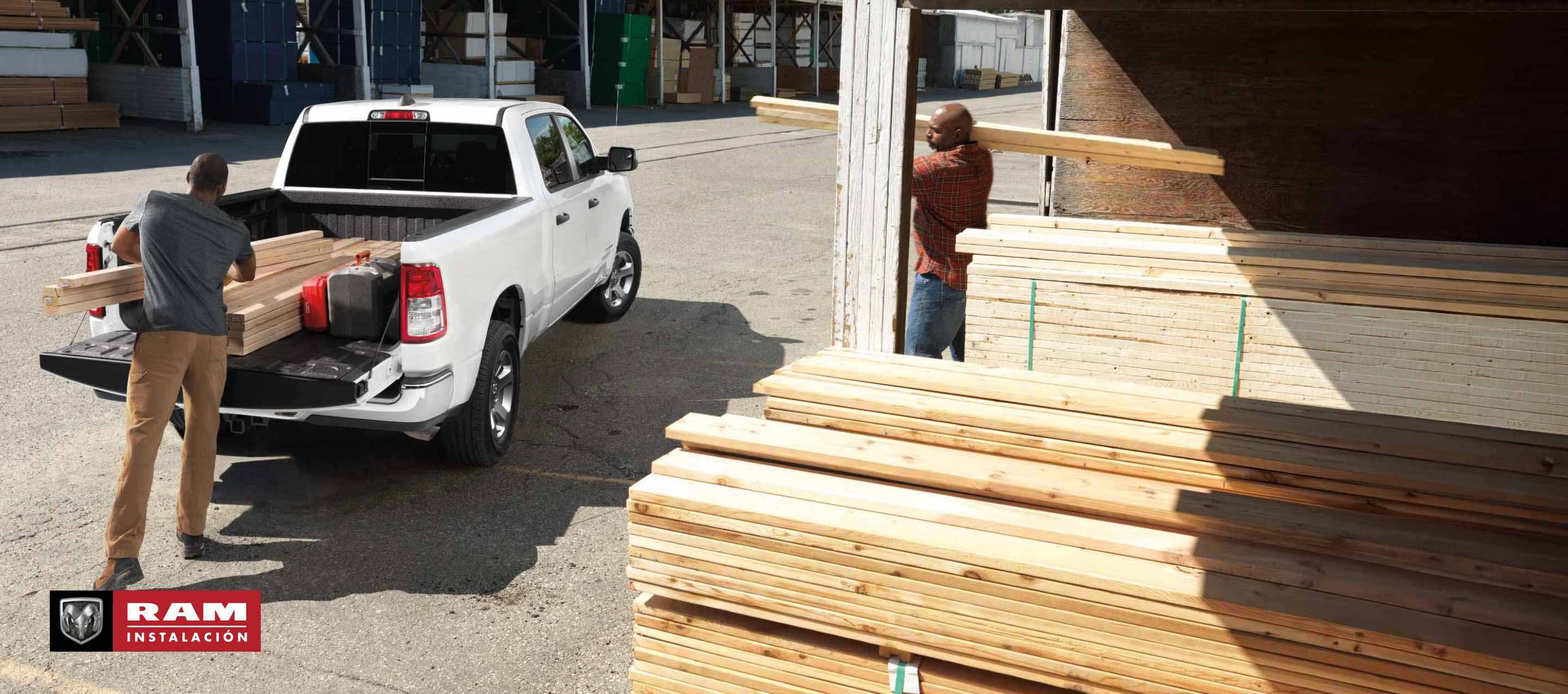 Vista trasera de una Ram 1500 Tradesman Crew Cab blanca 2024 estacionada en un depósito de madera con la puerta trasera abierta y un hombre cargando madera en la plataforma del camión. Ram Professional.