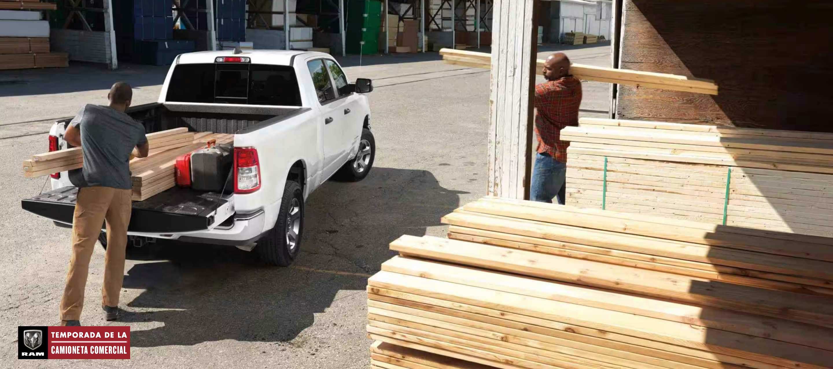Ángulo trasero de una Ram 1500 Tradesman Crew Cab 2024 blanca estacionada en un depósito de madera con la puerta trasera abierta y un hombre cargando madera en la plataforma de la camioneta mientras otro hombre trae más madera. Temporada de la camioneta comercial Ram.