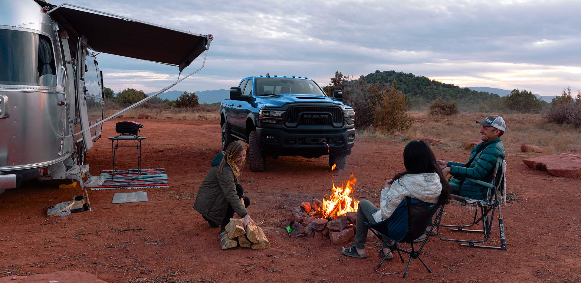 Se muestra una camioneta Ram 2500 Power Wagon Crew Cab 2024 azul estacionada en un campamento en el desierto, junto a una casa rodante y tres personas sentadas alrededor de un fogón.