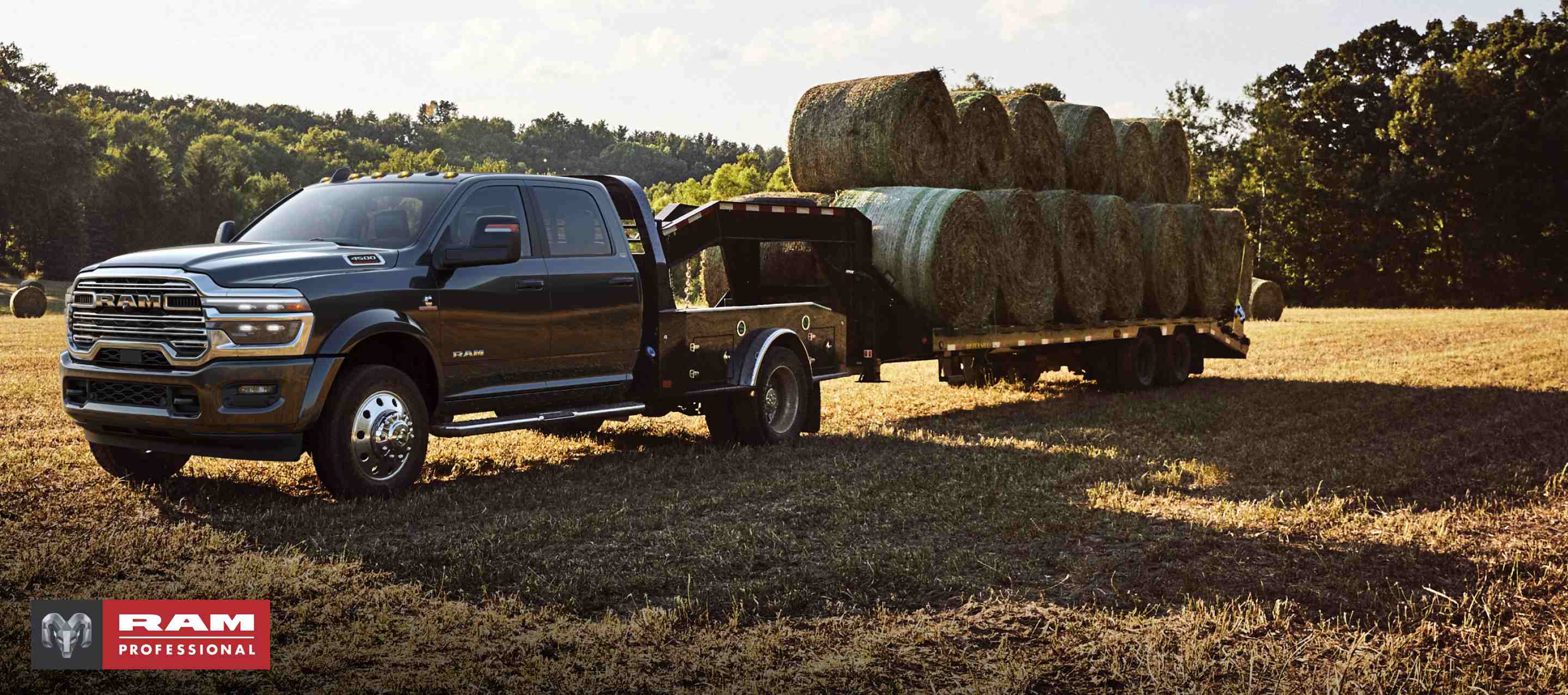 Una Ram 5500 Limited 4x4 Chassis Crew Cab 2024 negra con un remolque de cuello de cisne cargado con varios fardos de heno, estacionado en un campo abierto. Ram Professional.