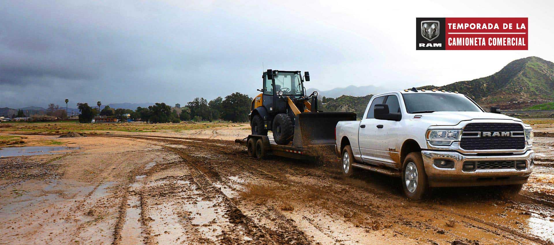 Una Ram 2500 Big Horn Crew Cab 2024 blanca remolca una pala mecánica sobre un tráiler de plataforma hacia un sitio de construcción lodoso. Temporada de camionetas comerciales.