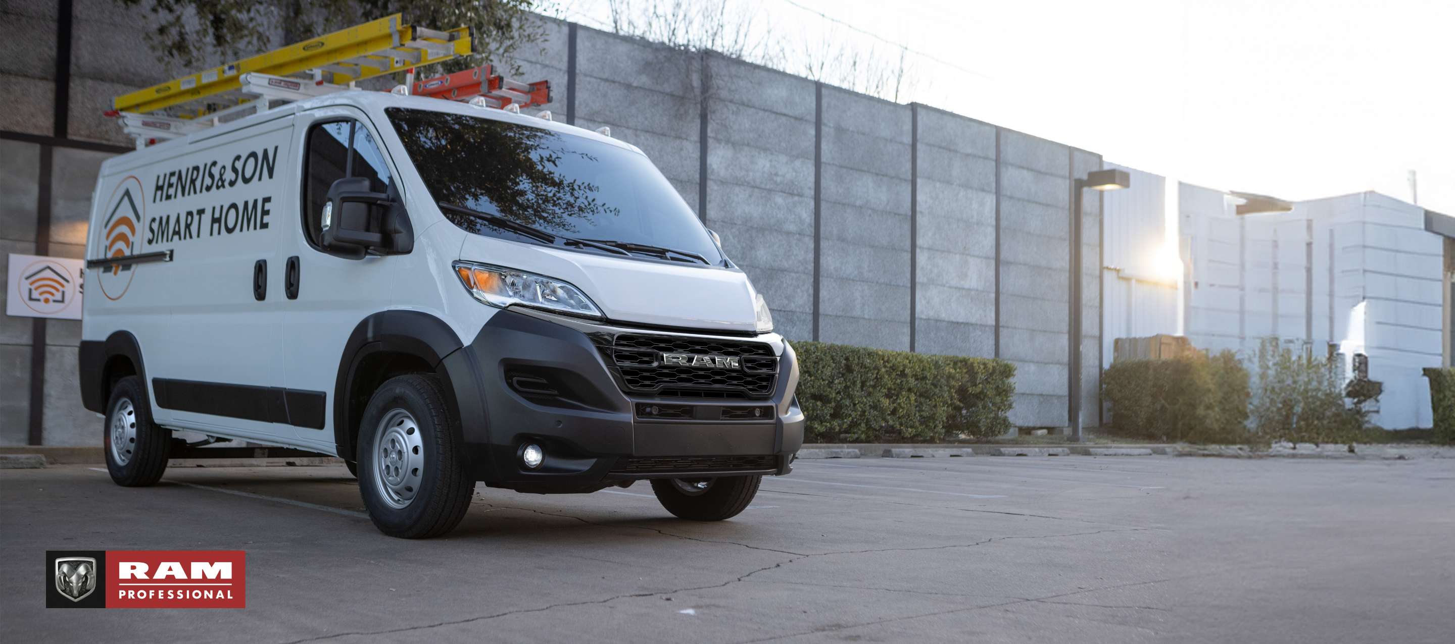 Una Ram ProMaster 1500 Tradesman Standard Roof Cargo Van 2024 blanca con dos escaleras en la parrilla portaequipaje y el logotipo de una compañía de instalación de hogares inteligentes en el panel lateral, estacionada al lado de un edificio comercial. Ram Professional.