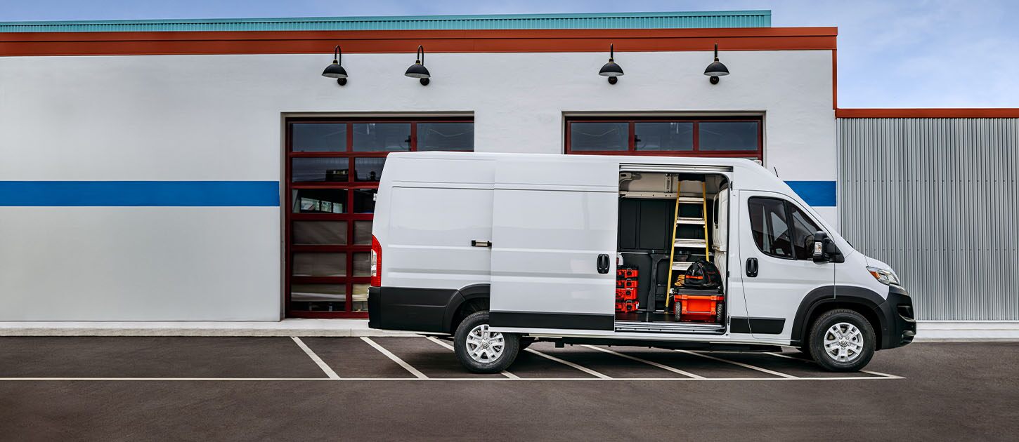 Perfil del lado del pasajero de una Ram ProMaster EV 3500 High Roof Cargo Van blanca estacionada al lado de un garaje comercial con su puerta lateral corrediza abierta, para revelar herramientas, equipos y una escalera en el interior.