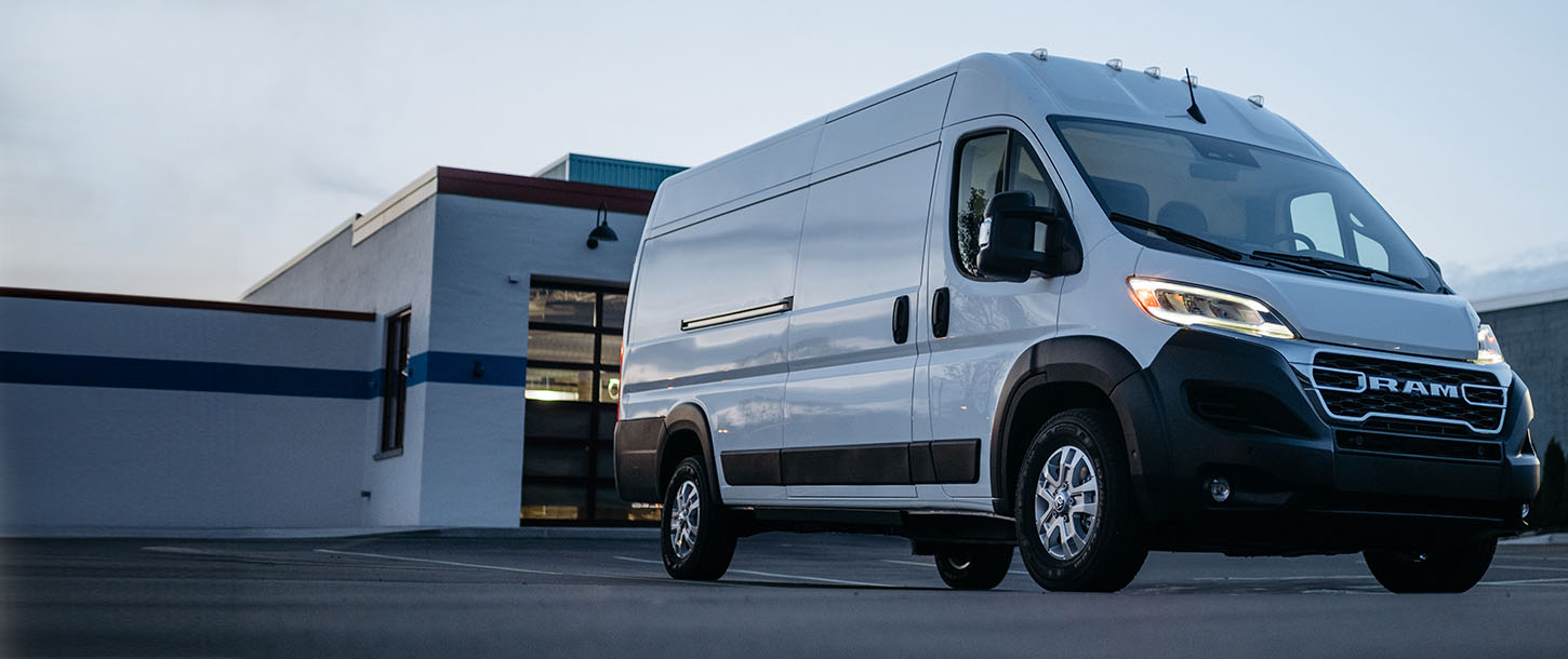 Ángulo delantero del lado del pasajero de una Ram ProMaster EV 3500 SLT High Roof Cargo Van blanca con los faros delanteros encendidos, estacionada en el estacionamiento de un edificio comercial.