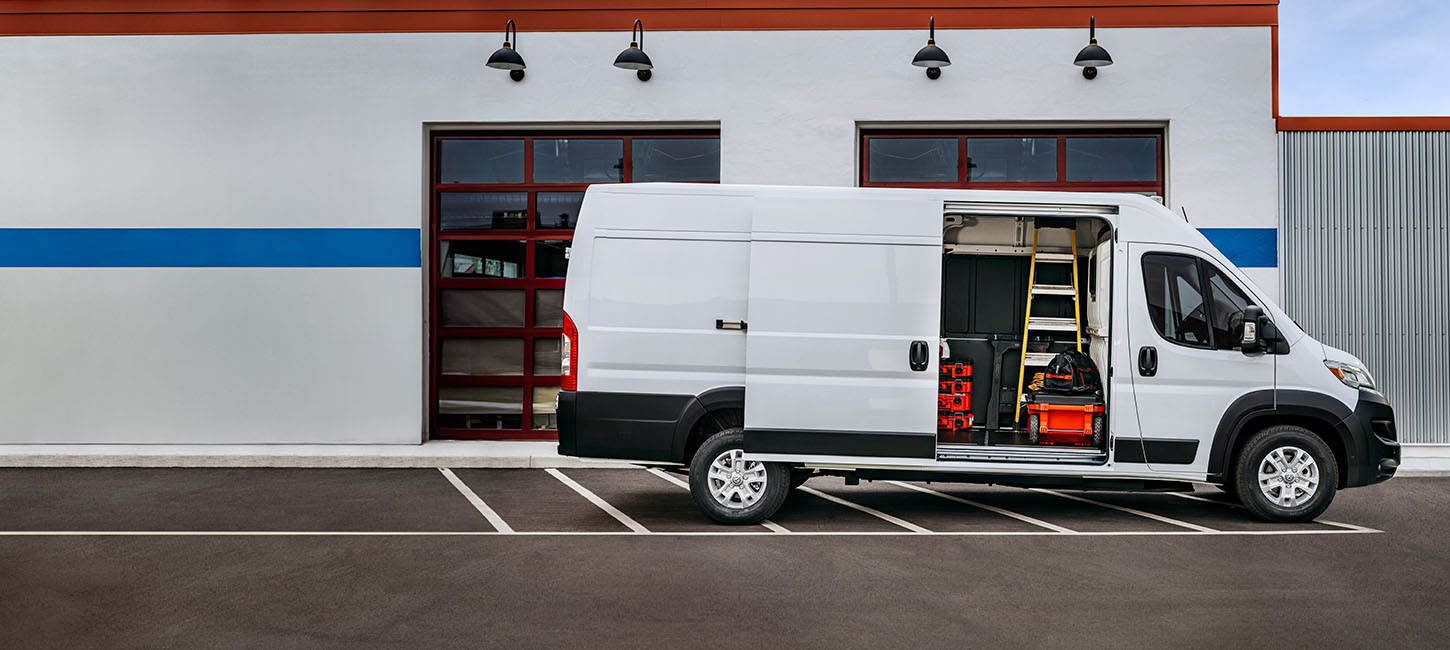 Perfil del lado del pasajero de una Ram ProMaster EV 3500 SLT High Roof Cargo Van blanca estacionada al lado de un garaje comercial con la puerta lateral corrediza abierta, lo que permite ver herramienta, equipo y una escalera en el interior.