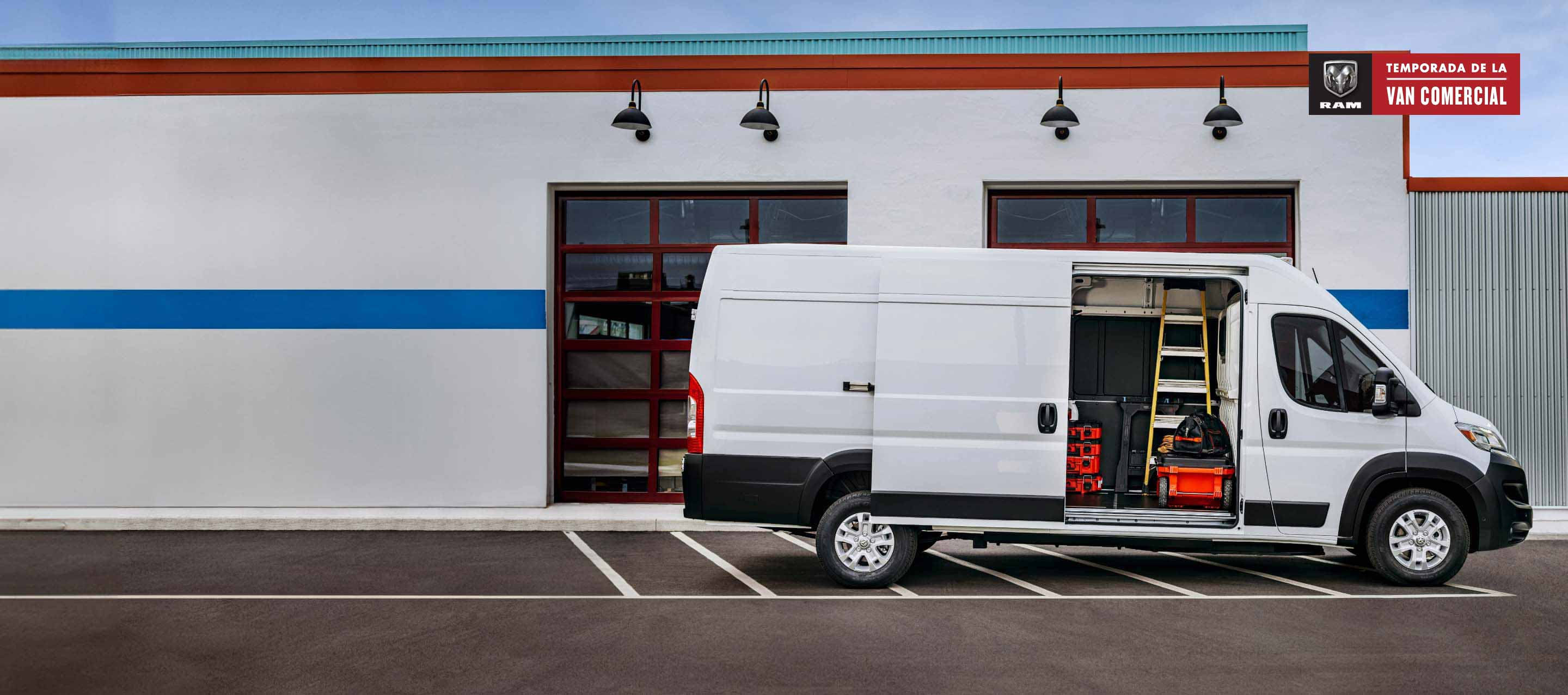 Perfil del lado del pasajero de una Ram ProMaster EV High Roof Cargo Van 2024 blanca (se muestra la Cargo Van de preproducción. El vehículo de producción real y las características pueden variar), estacionada al lado de un garaje comercial con la puerta lateral corrediza del lado del pasajero abierta, para exhibir una escalera, herramientas y equipos en el interior. Temporada de vans comerciales de Ram.