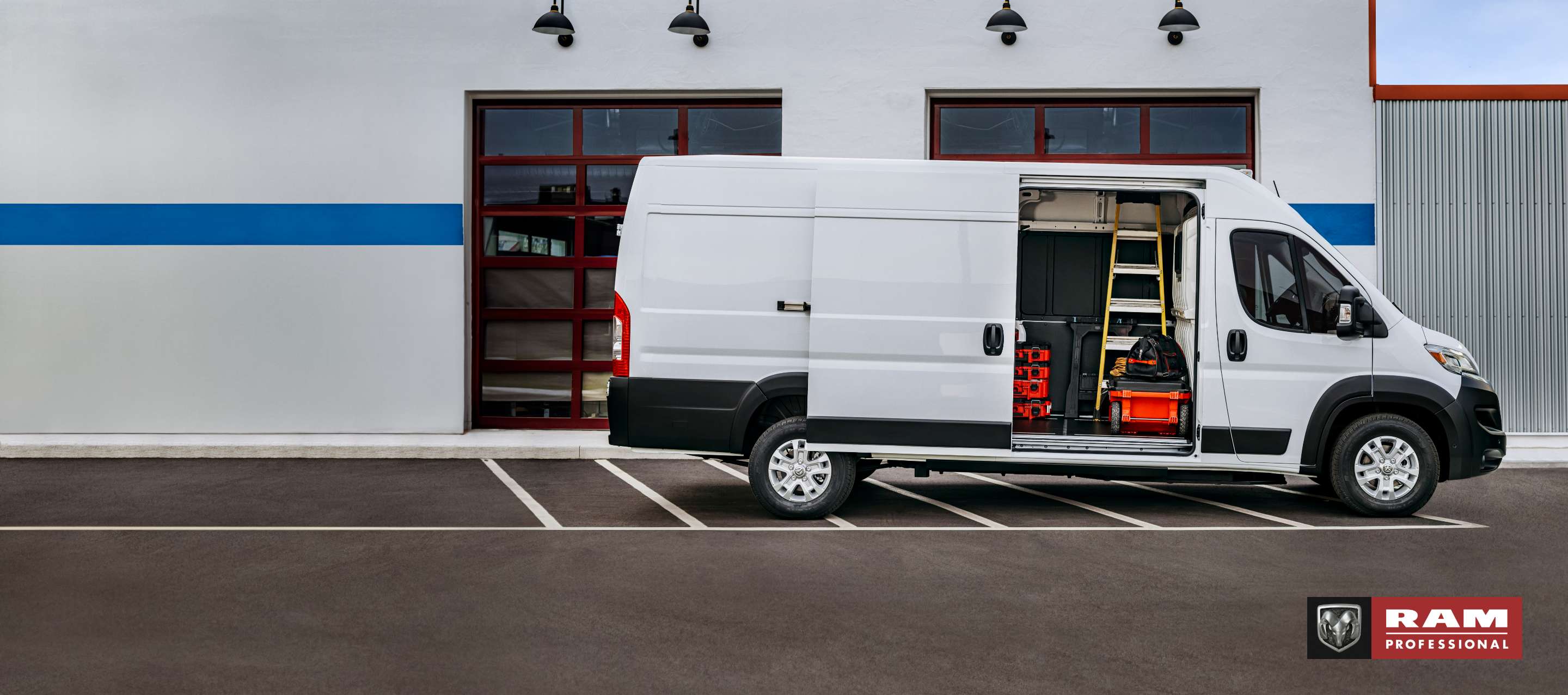 Perfil del lado del pasajero de una Ram ProMaster EV High Roof Cargo Van 2024 blanca (se muestra la Cargo Van de preproducción. El vehículo de producción real y las características pueden variar), estacionada al lado de un garaje comercial con la puerta lateral corrediza del lado del pasajero abierta, para exhibir una escalera, herramientas y equipos en el interior. Ram Professional.