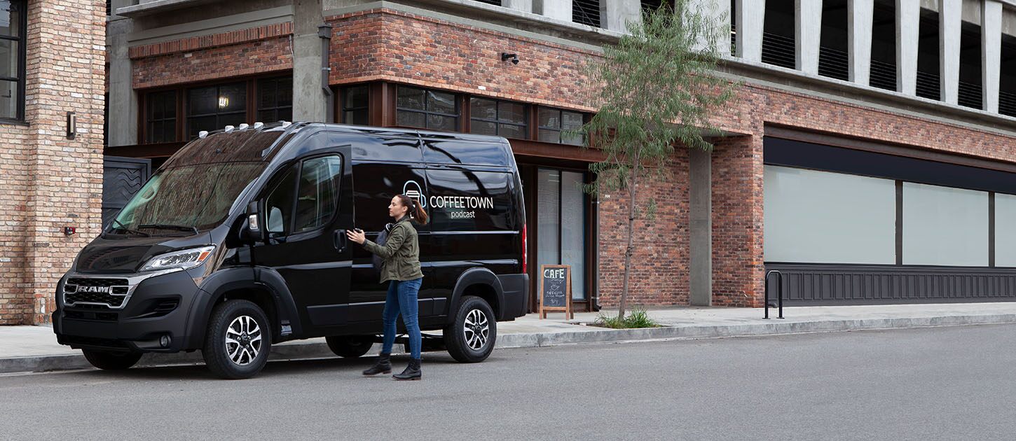 Una Ram ProMaster 2500 SLT Cargo Van High Roof 2024 negra con gráficas de una empresa de café en el panel trasero del lado del conductor, estacionada en una calle de la ciudad junto a una cafetería.