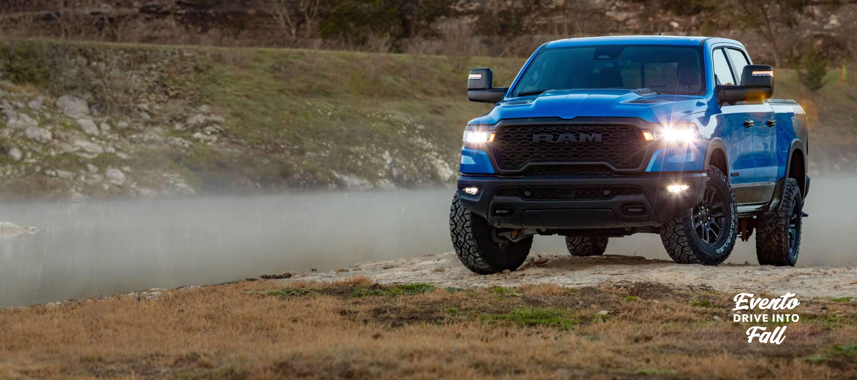 Una Ram 1500 Rebel 4x4 Crew Cab 2025 azul, con sus faros delanteros y antiniebla encendidos, sube una pequeña colina fuera de la carretera. Evento de ventas Drive Into Fall.