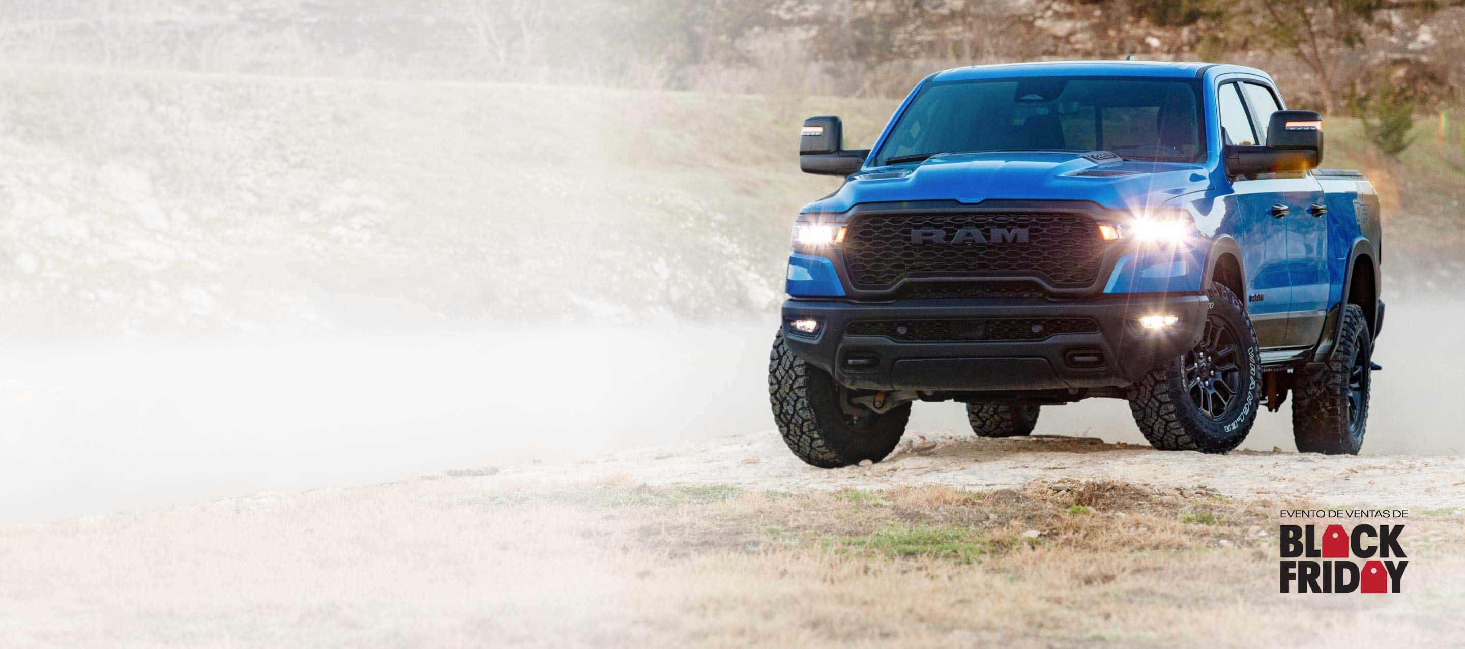 Una Ram 1500 Rebel 4x4 Crew Cab 2025 azul, con sus faros delanteros y antiniebla encendidos, sube una pequeña colina fuera de la carretera. Evento de ventas Black Friday.