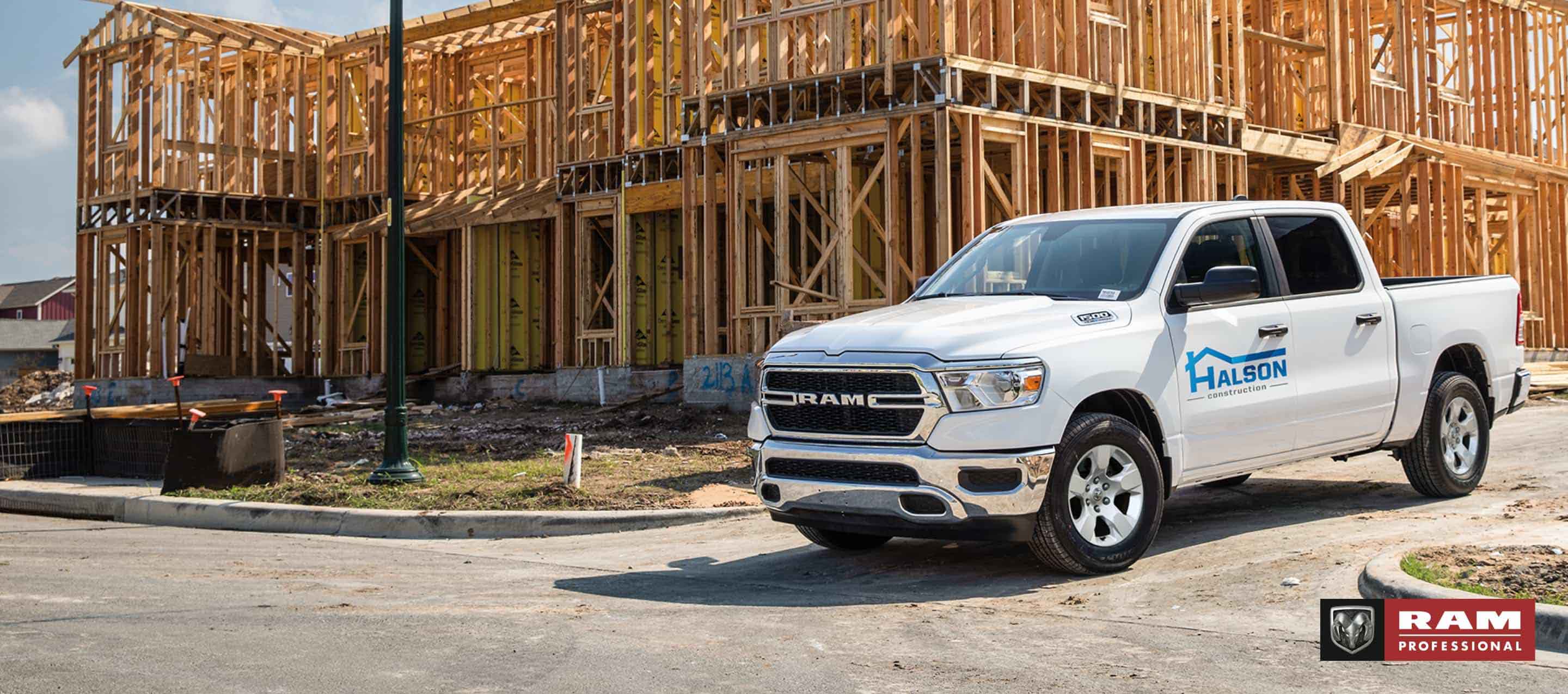 Una Ram 1500 Tradesman 4x4 Crew Cab 2025 blanca, con gráfico de una compañía de construcción en la puerta del conductor, saliendo de una obra de construcción comercial. Ram Professional.