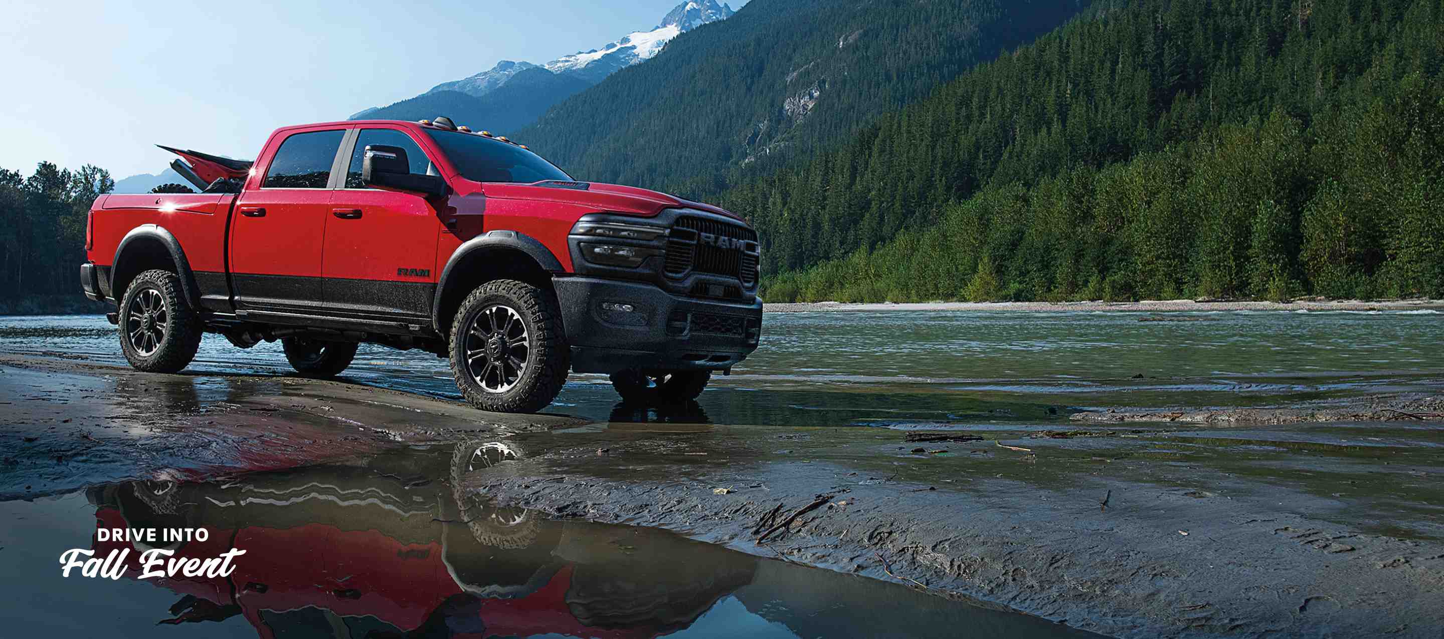 A red 2025 Ram 2500 Rebel 4x4 Crew Cab parked on a muddy trail beside a stream with mountains in the background. Evento de ventas Drive Into Fall.