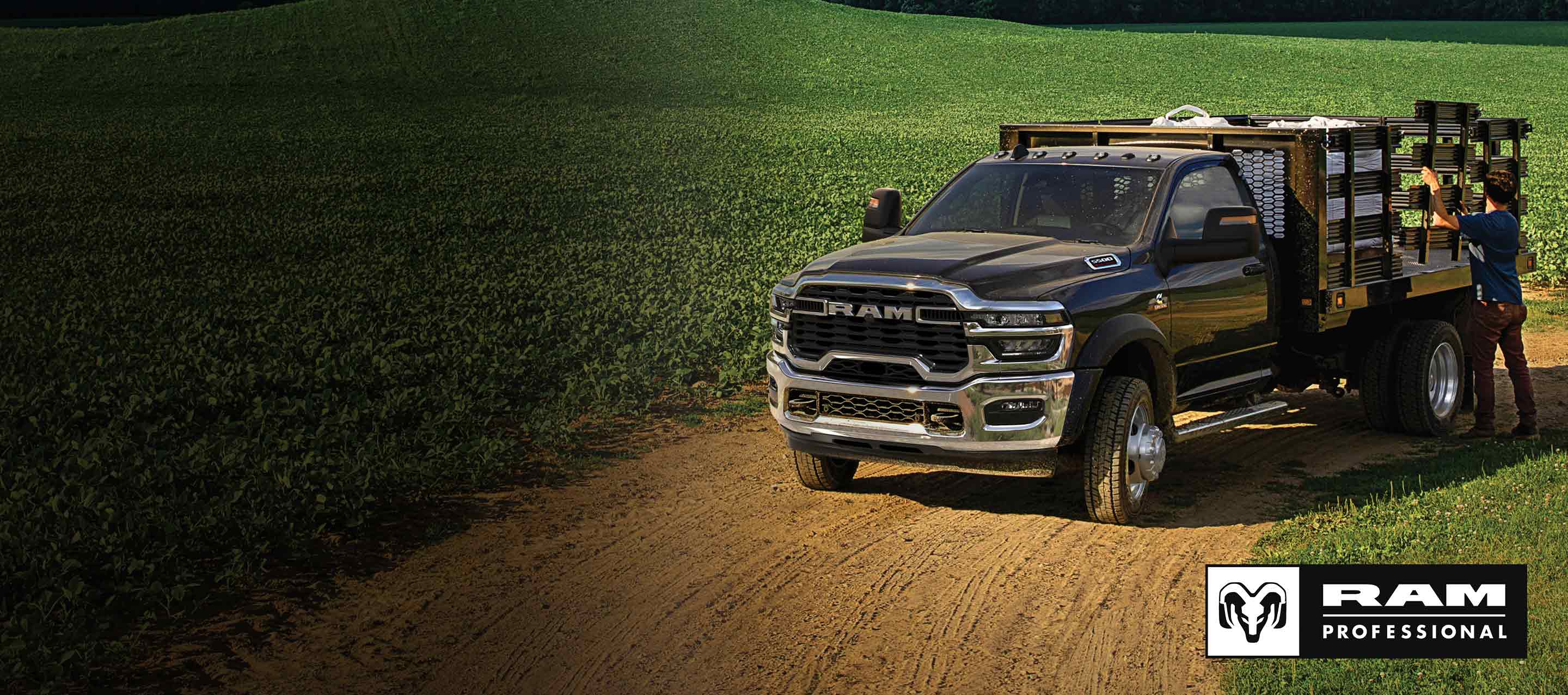 Una Ram 5500 Tradesman Chassis Cab Regular Cab 2025 negra con una mejora de paisajismo, estacionada en el gran campo de un granjero con un hombre ajustando uno de los paneles de mejora laterales. Ram Professional.