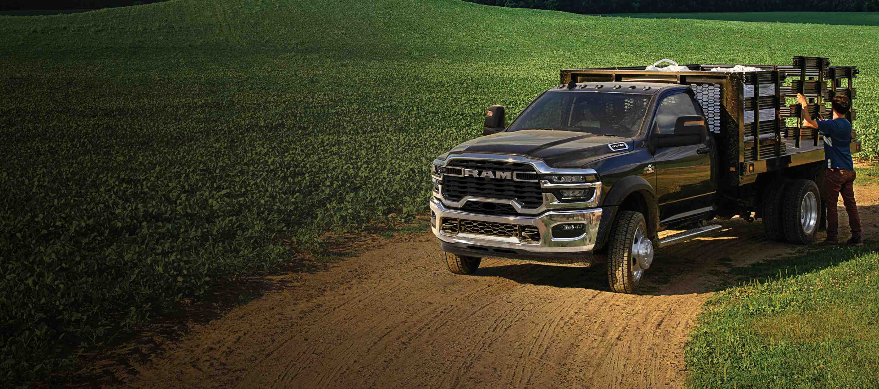 Una Ram 5500 Tradesman Chassis Cab Regular Cab 2025 negra con una mejora de paisajismo, estacionada en el gran campo de un granjero con un hombre ajustando uno de los paneles de mejora laterales. Ram Professional.