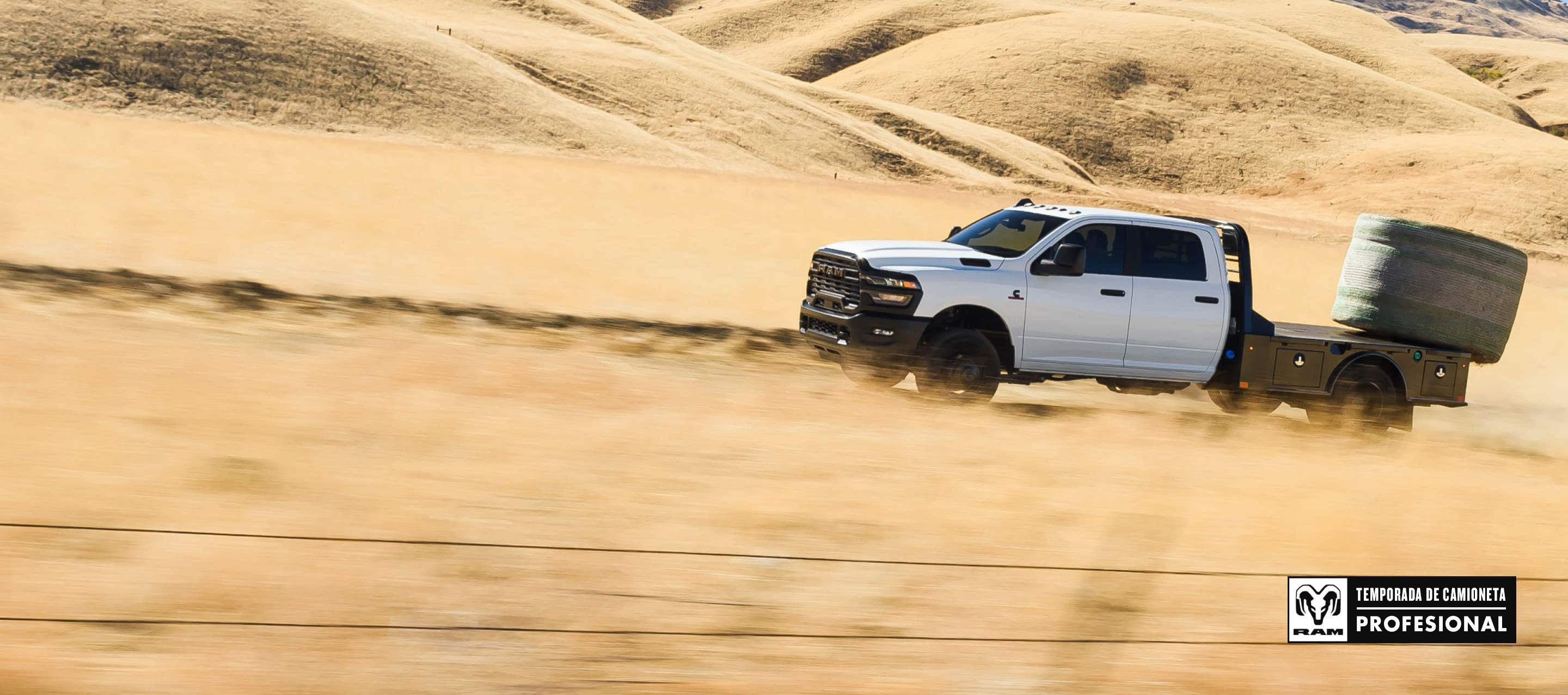 Una Ram 3500 Tradesman Crew Cab Chassis Cab 2025 blanca subiendo por una duna de arena en el desierto, con un enorme fardo de heno sobre su caja tipo plataforma. Temporada de camionetas Ram Professional.