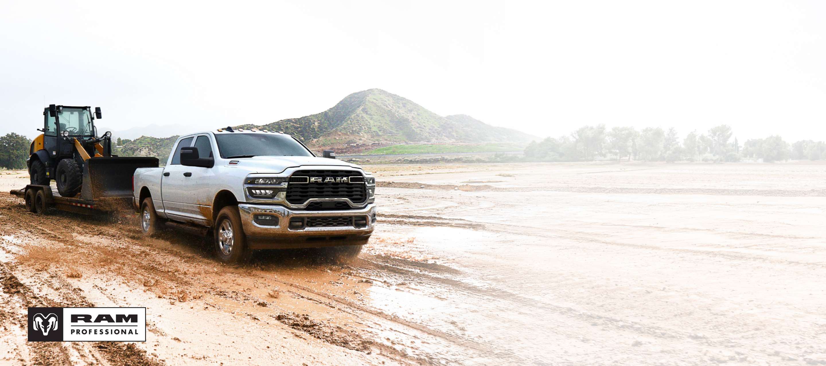 Una Ram 2500 Tradesman 4x4 Crew Cab 2025 blanca acarreando una pala mecánica sobre un remolque de plataforma plana por un sitio de construcción lodoso. Ram Professional.