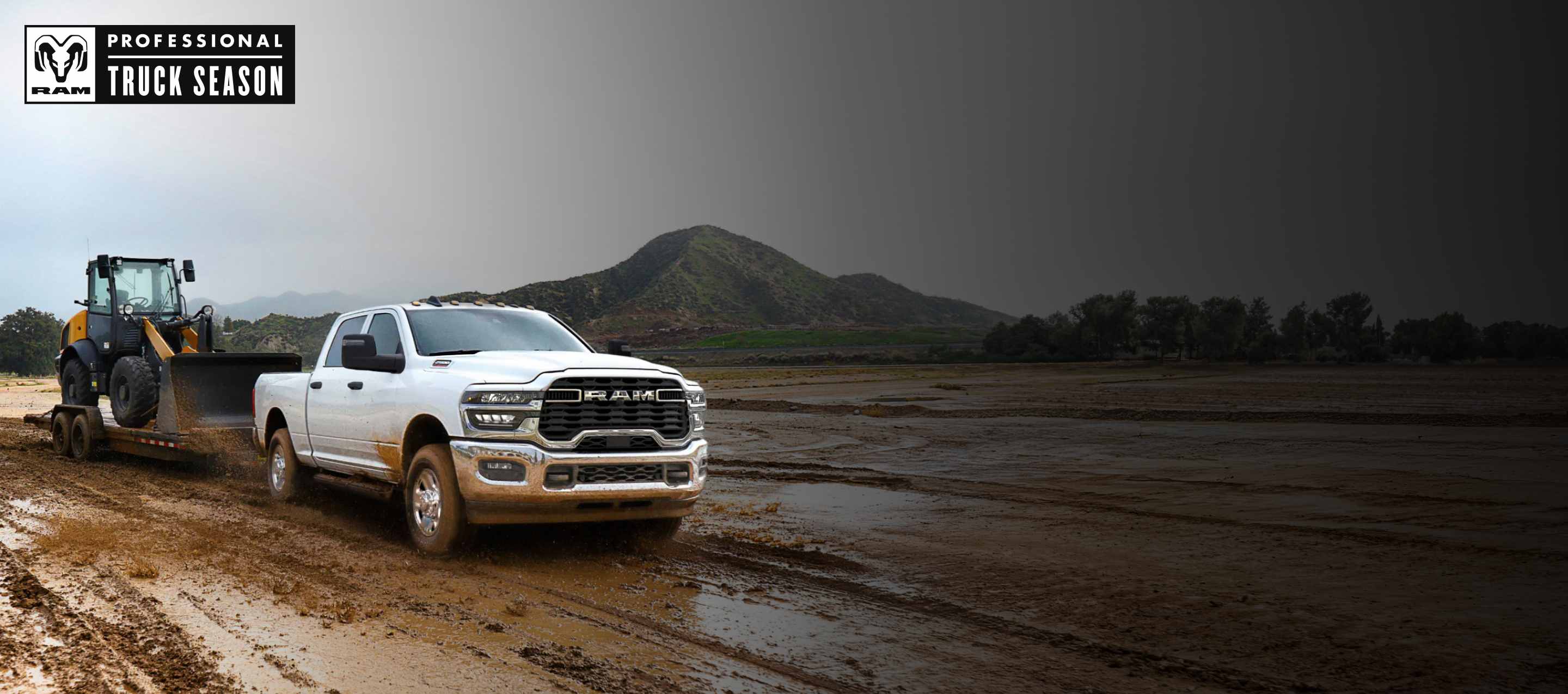 Una Ram 2500 Tradesman 4x4 Crew Cab 2025 blanca acarreando una pala mecánica sobre un remolque de plataforma plana por un sitio de construcción lodoso. Temporada de camionetas Ram Professional.