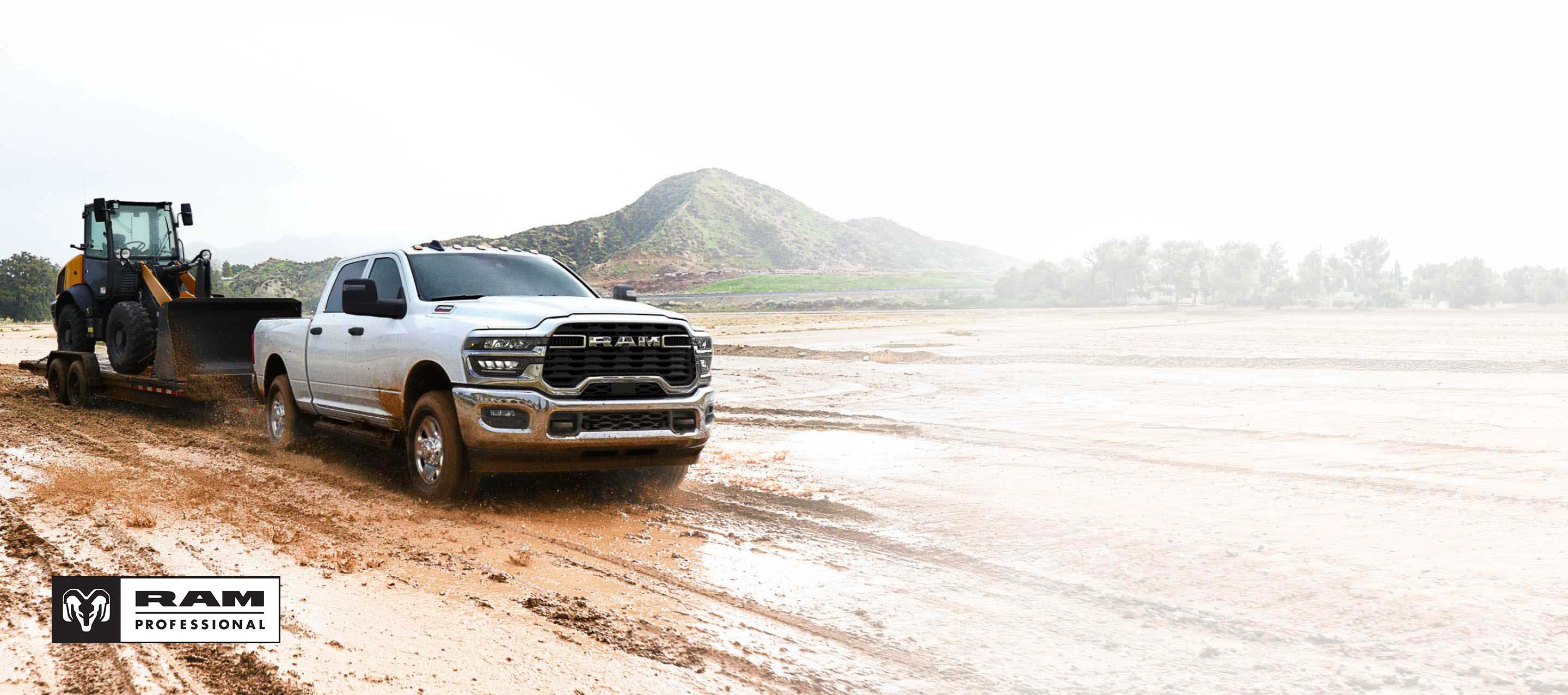 Una Ram 2500 Tradesman 4x4 Crew Cab 2025 blanca acarreando una pala mecánica sobre un remolque de plataforma plana por un sitio de construcción lodoso. Ram Professional.