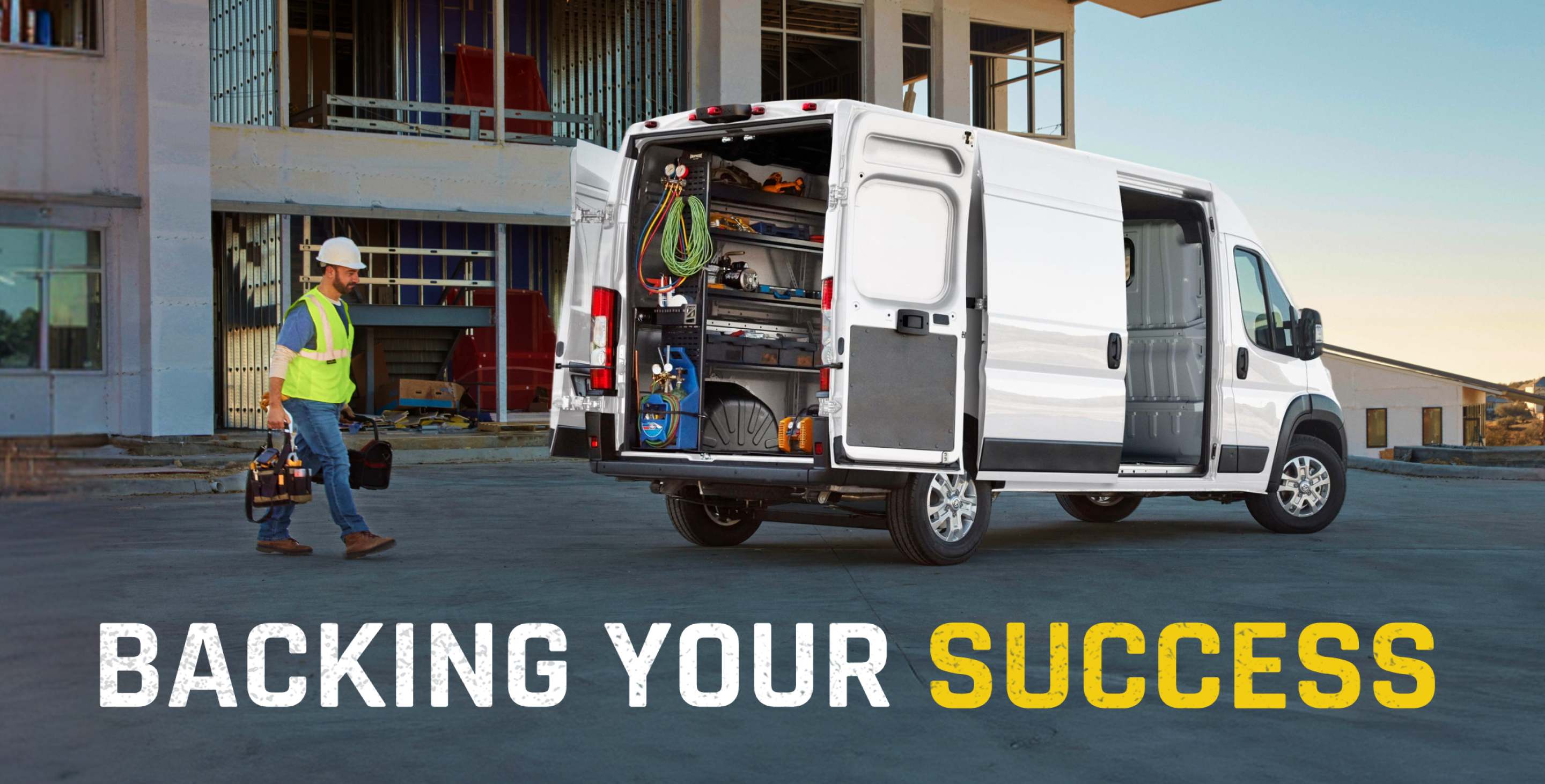 La parte trasera de una Ram ProMaster 3500 SLT Cargo Van High Roof 2026 blanca, con la puerta corrediza del lado del pasajero y las puertas traseras abiertas, mostrando estantes llenos de herramientas y un técnico caminando hacia la van.
