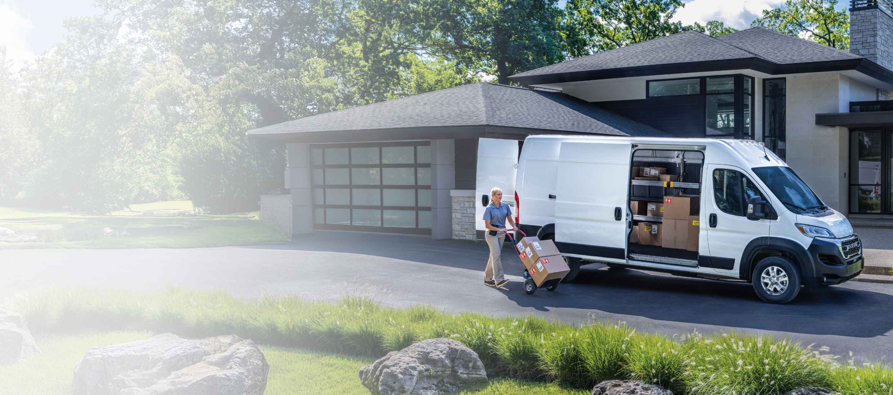 Una Cargo Van Ram ProMaster EV 3500 2026 SLT High Roof blanca con la puerta deslizable del lado del pasajero y las puertas traseras abiertas que permiten ver paquetes en el interior, estacionada en la entrada de una casa del vecindario, con un repartidor cerca empujando una carretilla llena de cajas.