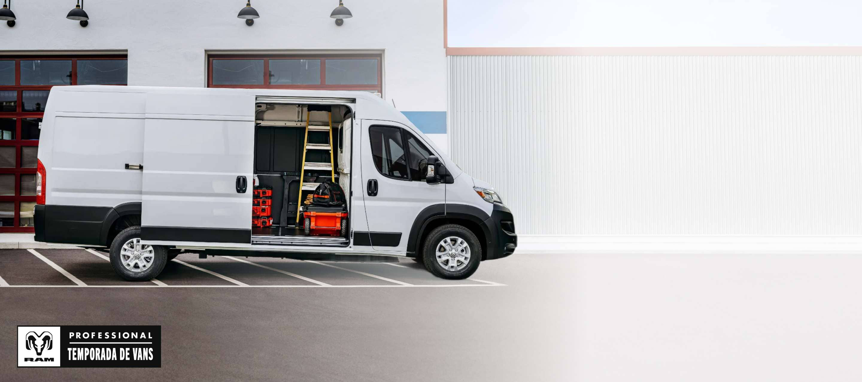 Perfil del lado del pasajero de una Ram ProMaster EV 3500 SLT High Roof Cargo Van 2025 blanca estacionada al lado de un garaje comercial con la puerta lateral corrediza abierta, lo que permite ver herramientas, equipos y una escalera en el interior. Temporada de vans profesionales Ram.