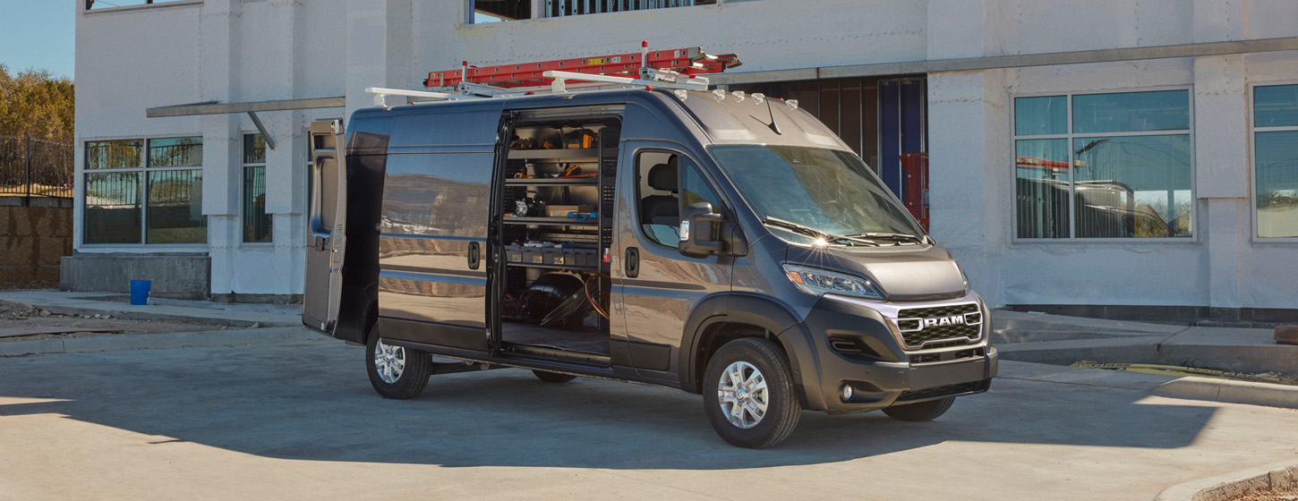 Un perfil del lado del pasajero de una Ram ProMaster 3500 SLT Plus Cargo Van High Roof 2025 gris estacionada al lado de un edificio comercial con una escalera en la parrilla portaequipaje y las puertas traseras y la puerta lateral deslizante abiertas para revelar estantes llenos de herramientas en la zona de carga.