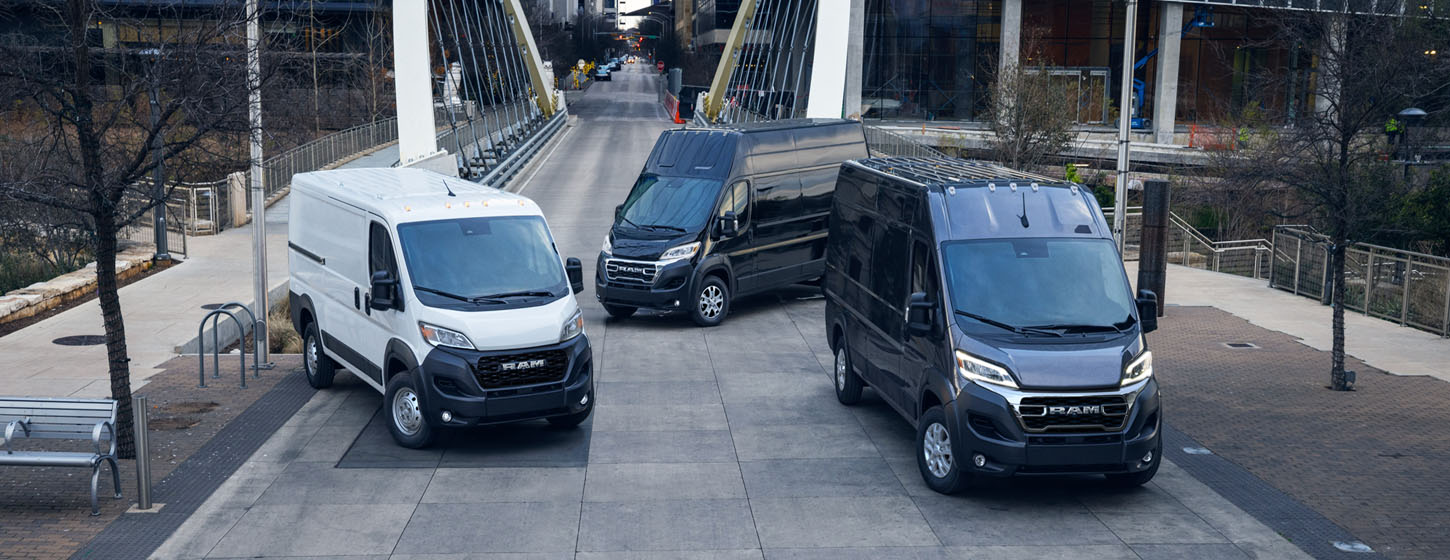 Tres modelos de Ram ProMaster 2025 estacionados al otro lado de un puente, en el centro de una gran ciudad, de izquierda a derecha: una Ram ProMaster 1500 Tradesman Cargo Van techo estándar blanca, una Ram ProMaster 3500 SLT Plus Cargo Van Super High Roof negra y una Ram ProMaster 3500 SLT Cargo Van High Roof negra.