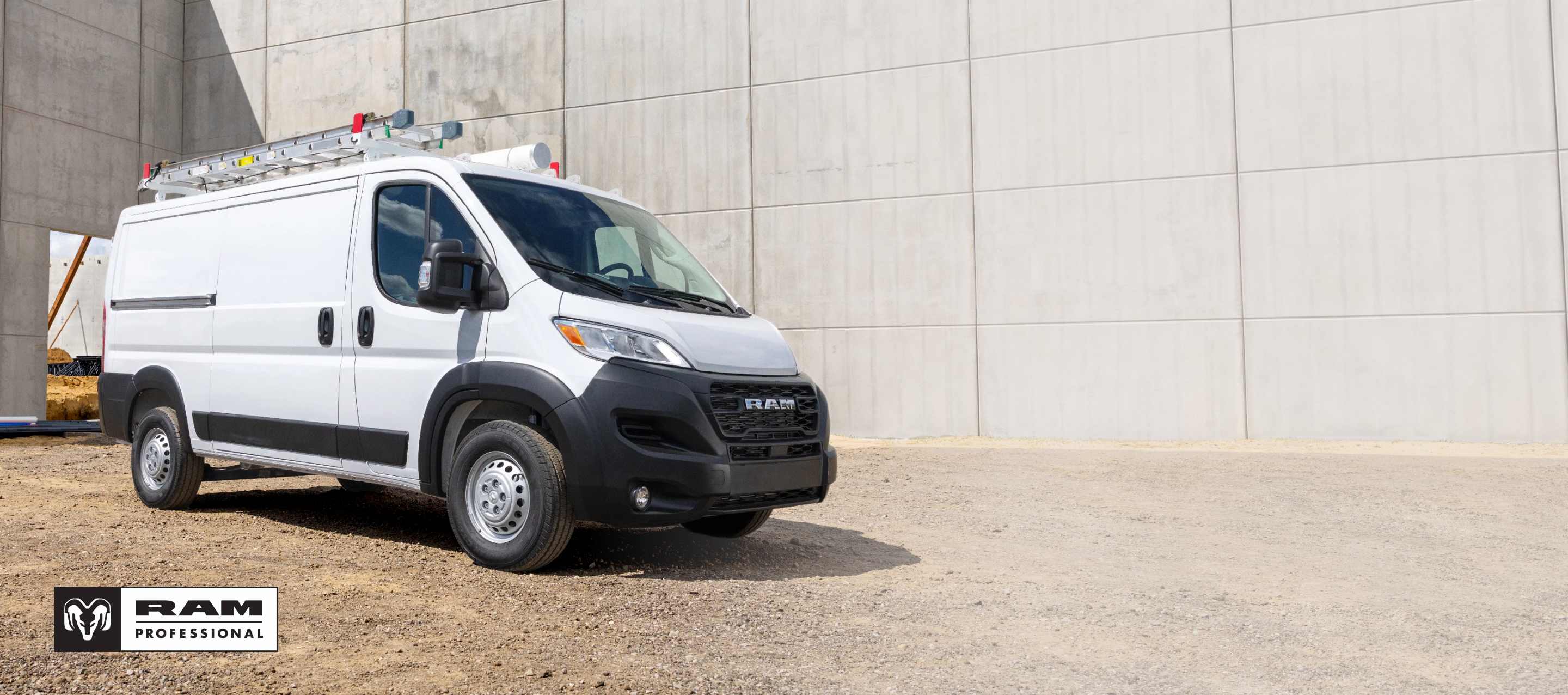 Ángulo delantero del lado del pasajero de una Ram ProMaster 1500 Tradesman Standard Roof Cargo Van 2025 blanca estacionada en una obra en construcción. Ram Professional.
