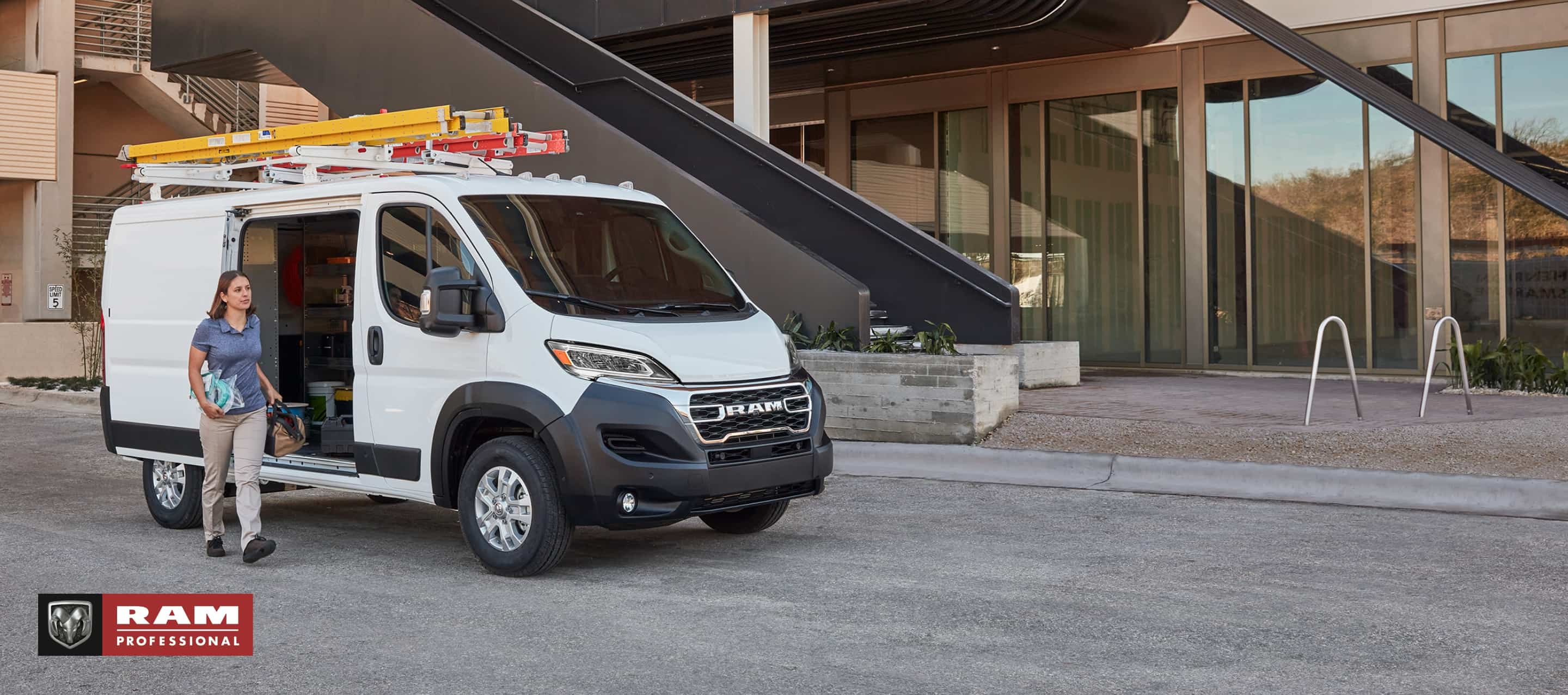 Ángulo delantero del lado del pasajero de una Ram ProMaster SLT Standard Roof Cargo Van 2025 blanca, estacionada al lado de un edificio moderno, con dos escaleras en la parrilla portaequipaje y la puerta lateral corrediza del lado del pasajero abierta. Ram Professional.