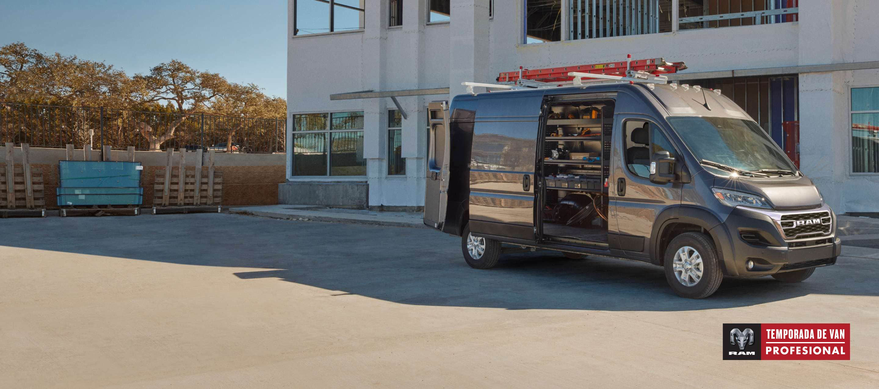 Un perfil del lado del pasajero de una Ram ProMaster 3500 SLT Plus High Roof Cargo Van 2025 gris estacionada al lado de un edificio comercial con una escalera en su parrilla portaequipaje, sus puertas traseras y puerta lateral corrediza abiertas, lo que permite visualizar estanterías llenas de herramientas en el área de carga. Temporada de vans profesionales.