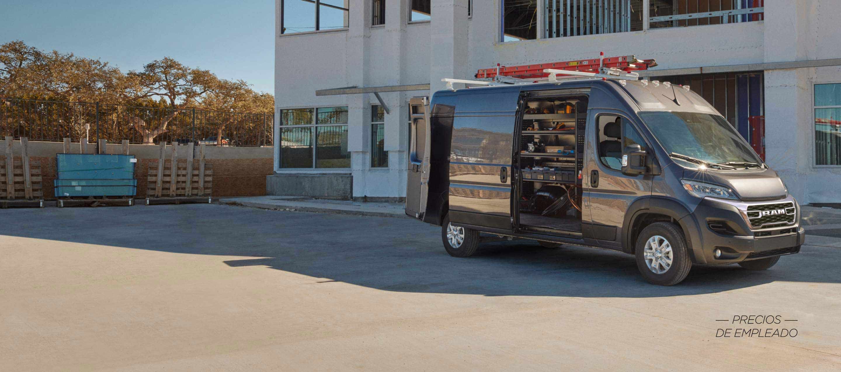 Un perfil del lado del pasajero de una Ram ProMaster 3500 SLT Plus High Roof Cargo Van 2025 gris estacionada al lado de un edificio comercial con una escalera en su parrilla portaequipaje, sus puertas traseras y puerta lateral corrediza abiertas, lo que permite visualizar estanterías llenas de herramientas en el área de carga. Evento de ventas Precios de empleado.