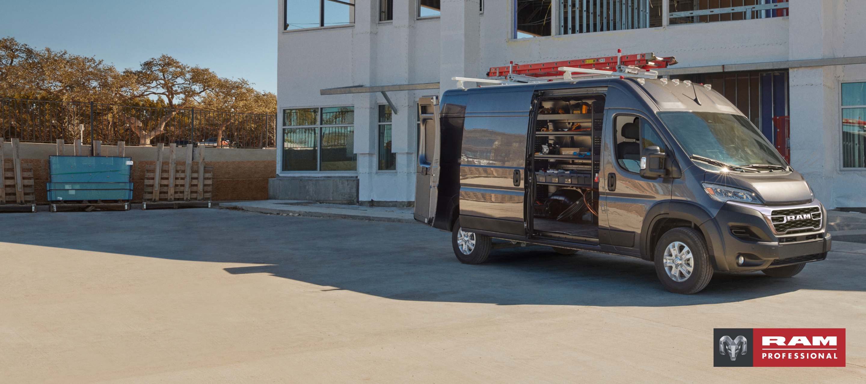 Un perfil del lado del pasajero de una Ram ProMaster 3500 SLT Plus High Roof Cargo Van 2025 gris estacionada al lado de un edificio comercial con una escalera en su parrilla portaequipaje, sus puertas traseras y puerta lateral corrediza abiertas, lo que permite visualizar estanterías llenas de herramientas en el área de carga. Ram Professional.