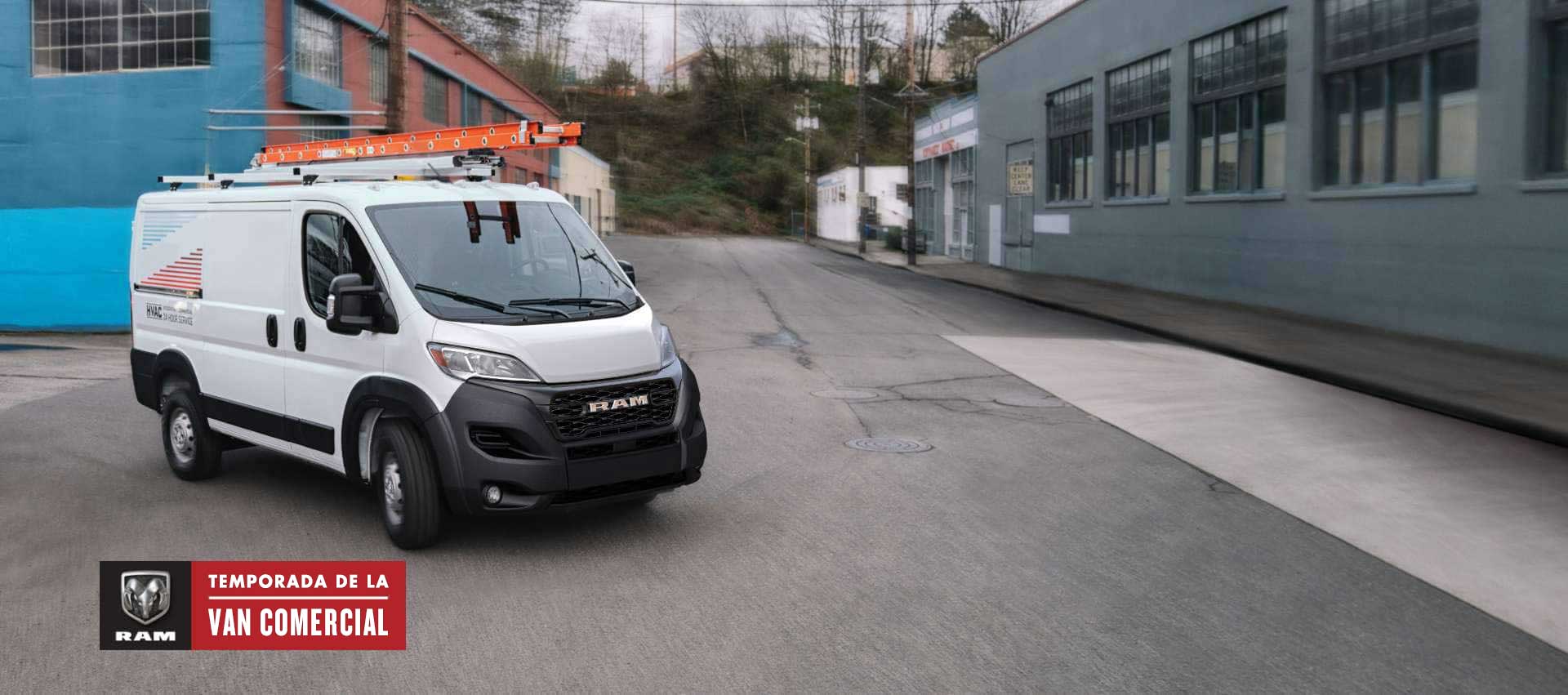 Ángulo delantero del lado del pasajero de una Ram ProMaster SLT Standard Roof Cargo Van 2025 blanca, estacionada al lado de un edificio moderno, con dos escaleras en la parrilla portaequipaje y la puerta lateral corrediza del lado del pasajero abierta. Ram Professional.