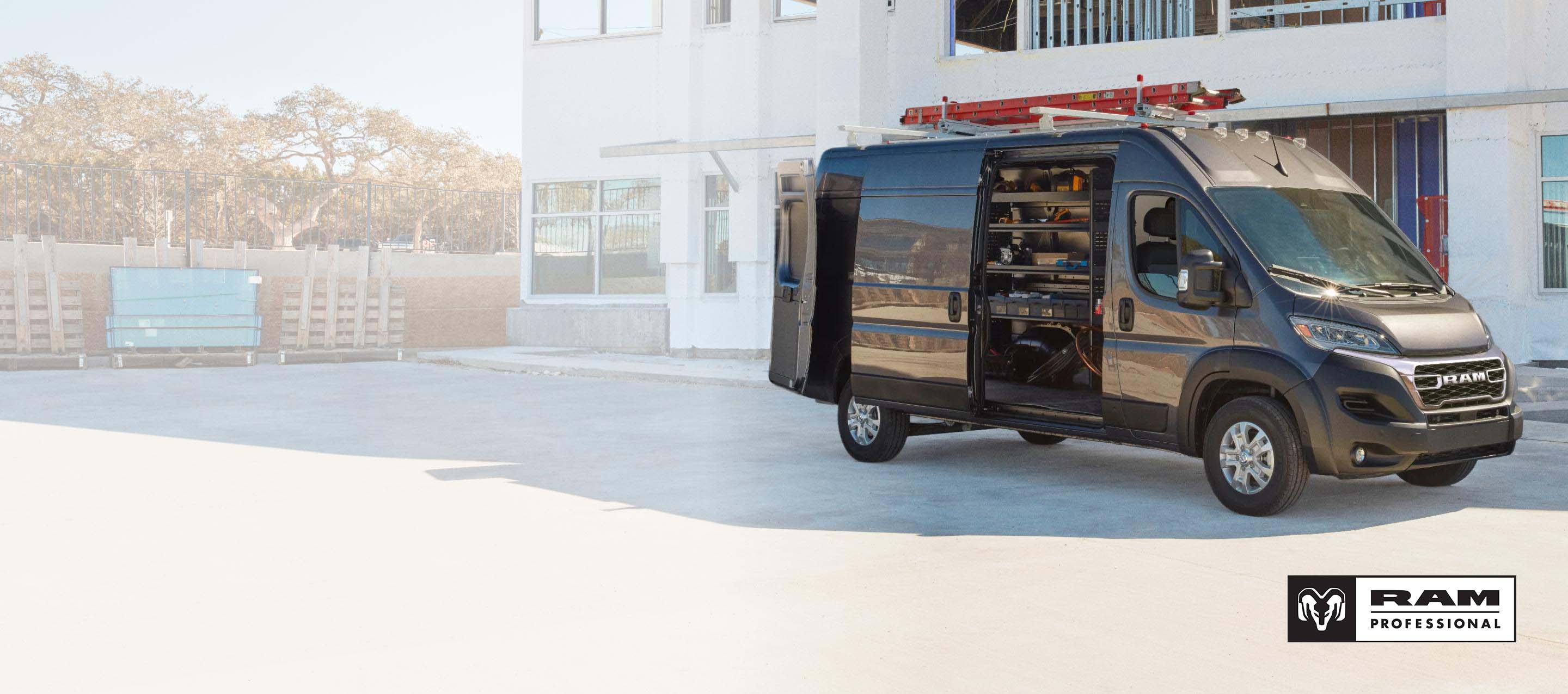 Una Ram ProMaster 3500 SLT High Roof Cargo Van 2025 gris estacionada al lado de un edificio comercial, con una escalera amarrada a la parrilla portaequipajes, y las puertas traseras y la puerta lateral corrediza abiertas para exhibir estanterías llenas de herramientas. Ram Professional.