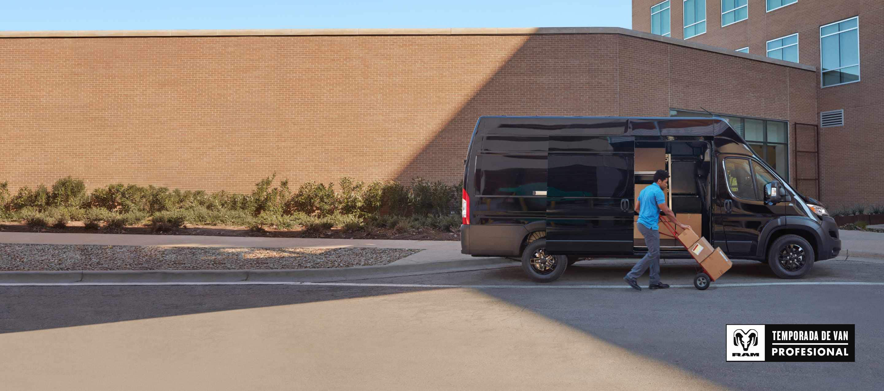 Un hombre empuja paquetes sobre un carrito cerca de la puerta corrediza del lado del pasajero de una cargo van Ram ProMaster 3500 SLT Plus Super High Roof 2025 negra con el paquete de apariencia negra. Temporada de vans profesionales Ram.