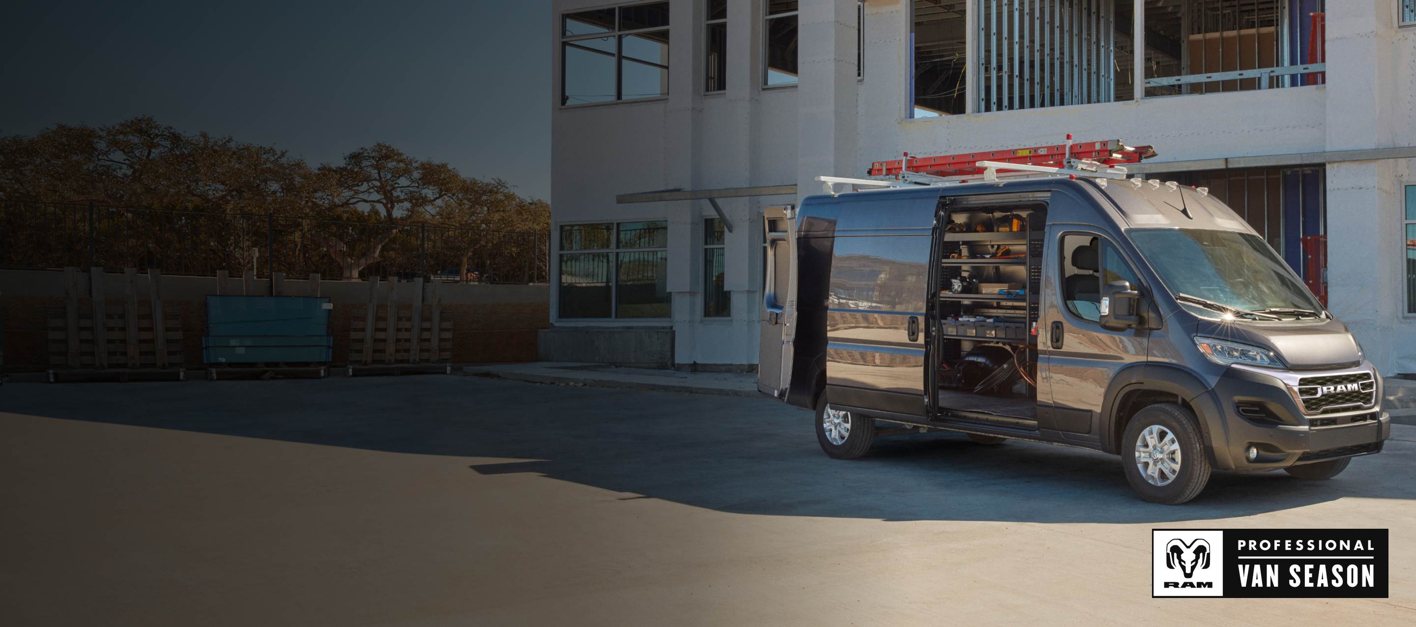 A dark gray 2025 Ram ProMaster 2500 SLT High Roof Cargo Van parked beside a commercial building with a ladder strapped to its roof rack, its rear doors and sliding side door open revealing shelving full of tools. Temporada de vans profesionales Ram.