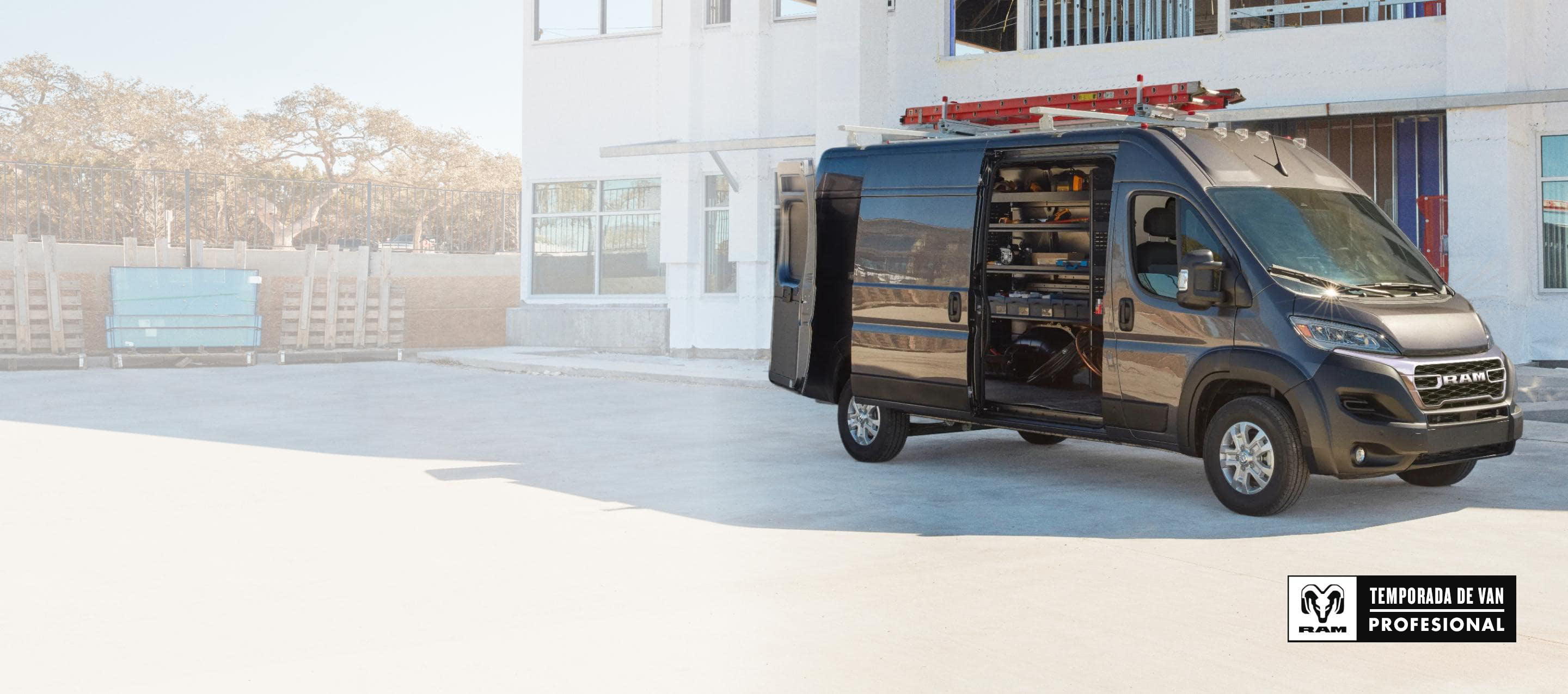 Una Ram ProMaster 3500 SLT High Roof Cargo Van 2025 gris estacionada al lado de un edificio comercial, con una escalera amarrada a la parrilla portaequipajes, y las puertas traseras y la puerta lateral corrediza abiertas para exhibir estanterías llenas de herramientas. Temporada de vans profesionales Ram.