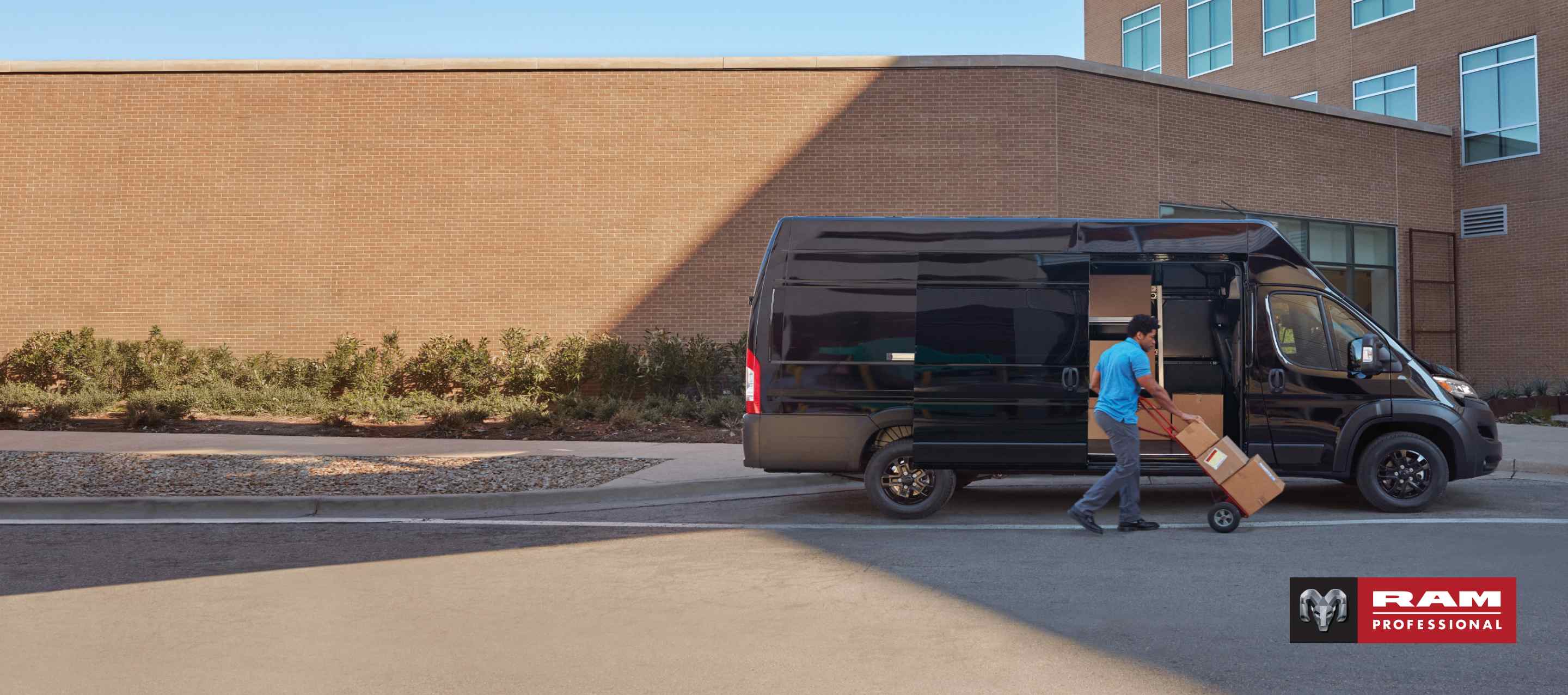 Un hombre empuja paquetes sobre un carrito cerca de la puerta corrediza del lado del pasajero de una cargo van Ram ProMaster 3500 SLT Plus Super High Roof 2025 negra con el paquete de apariencia negra. Ram Professional.