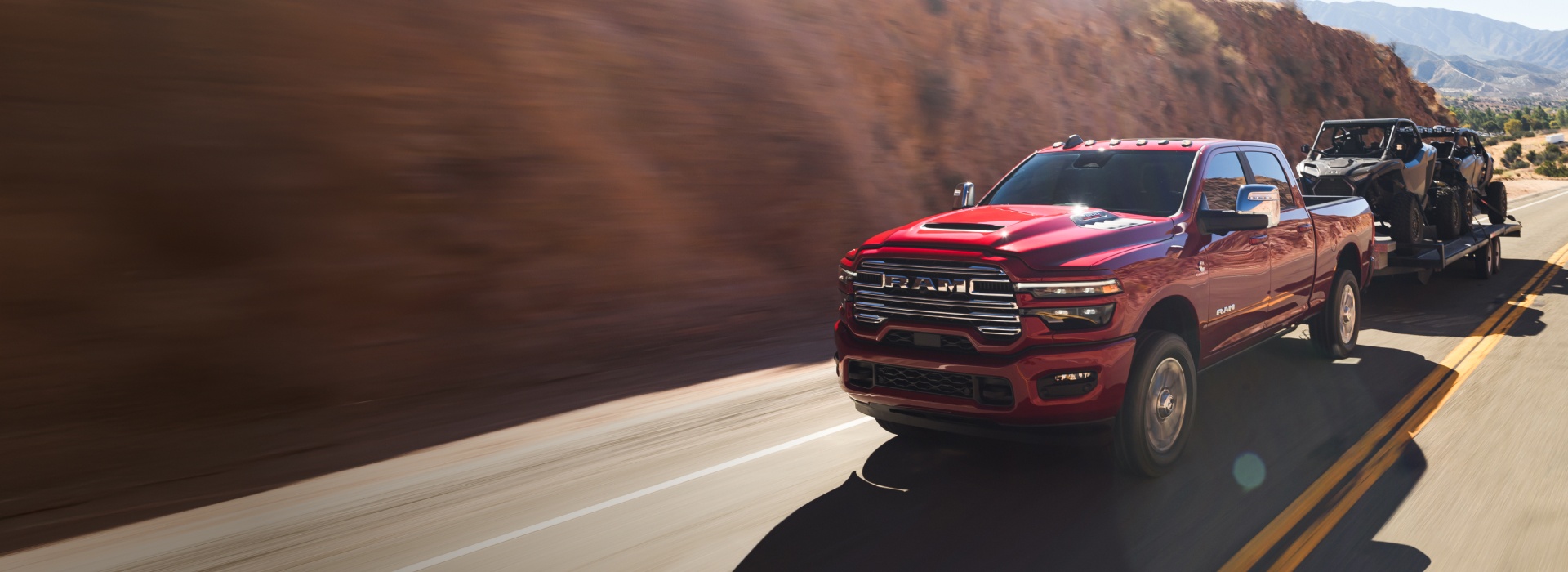 Una Ram 2500 Laramie 4x4 Crew Cab 2026 roja circulando por una carretera en las montañas, mientras remolca dos ATV.