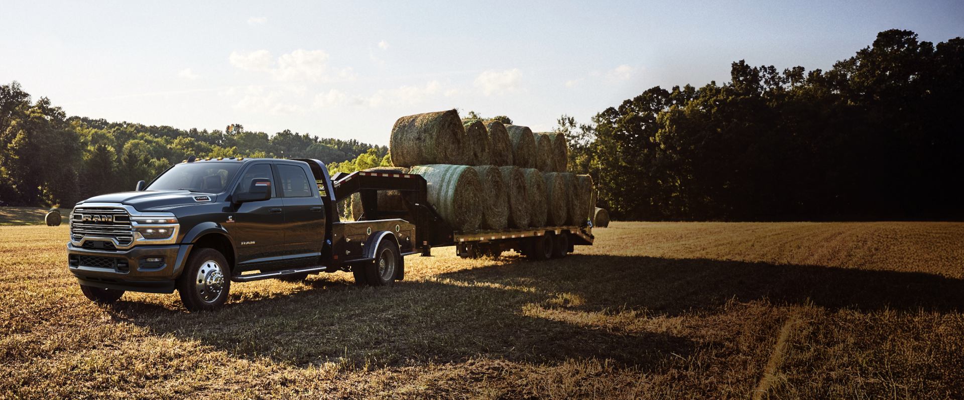 Una Ram 5500 Laramie Chassis Cab Crew Cab 2026 negra con un remolque tipo cuello de cisne cargada con varios fardos de heno, estacionada en un campo.