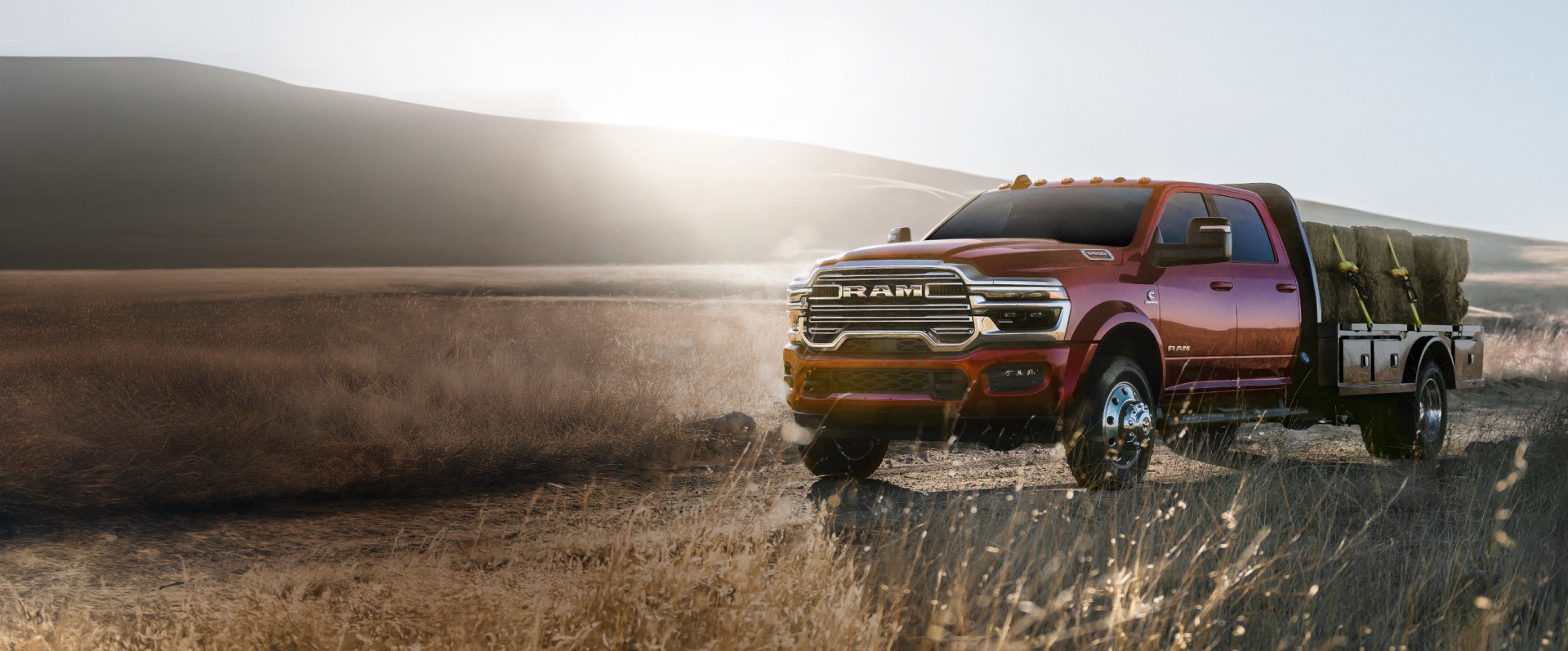 Una Ram 5500 Laramie Chassis Cab Crew Cab 2026 roja trasladando varios fardos de heno grandes en una mejora de plataforma larga mientras atraviesa tierras de ranchos.