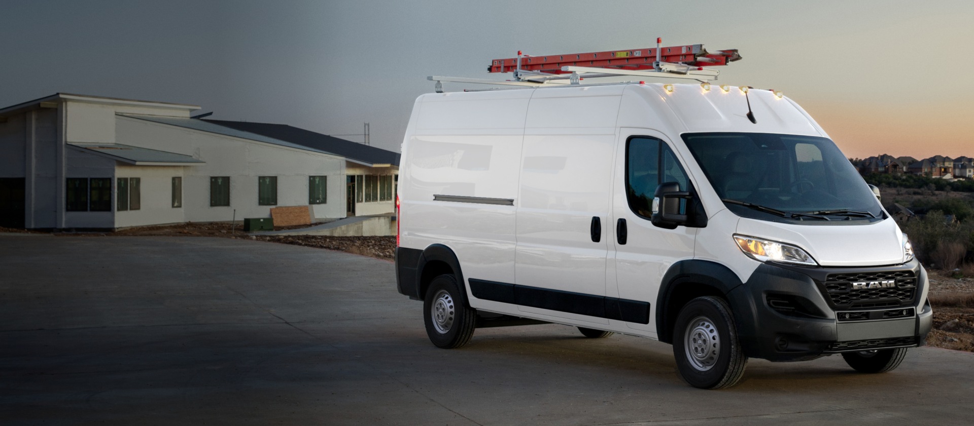 Ángulo delantero del lado del pasajero de una Ram ProMaster Tradesman High Roof Cargo Van 2026 blanca con una escalera en la parrilla portaequipaje, estacionada cerca de una obra.