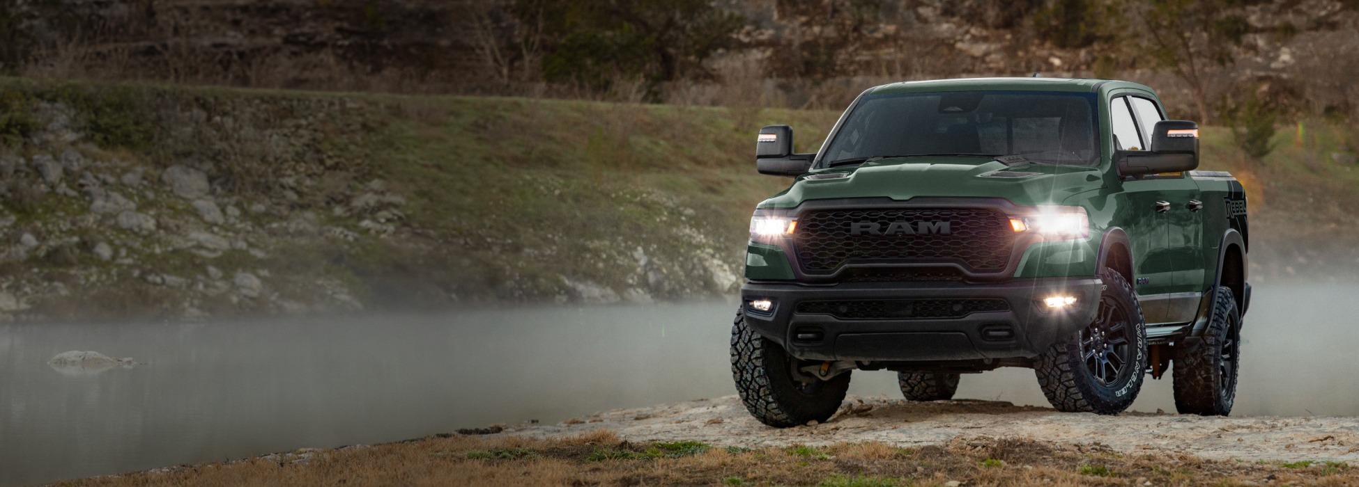 Una Ram 1500 Rebel 4x4 Crew Cab 2026 en color verde, con los faros delanteros encendidos, avanzando por un camino todoterreno.
