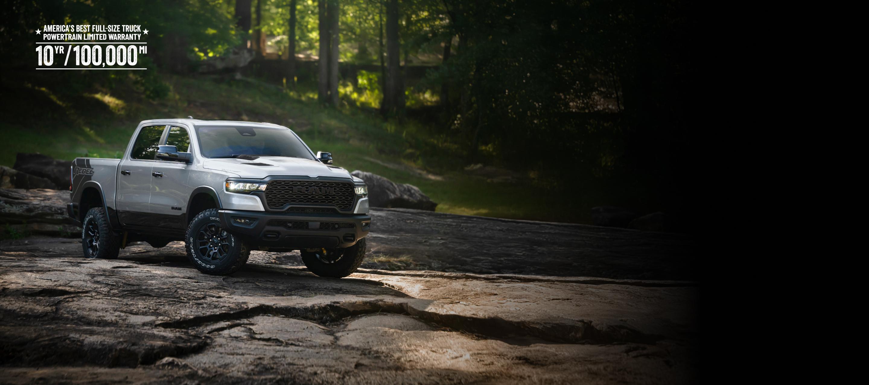 Una Ram 1500 Rebel 4x4 Crew Cab 2026 blanca circulando por un sendero rocoso fuera de la carretera, con un bosque de fondo. La mejor garantía limitada del tren motriz para camionetas de tamaño completo de los Estados Unidos. 10 años, 100,000 millas.