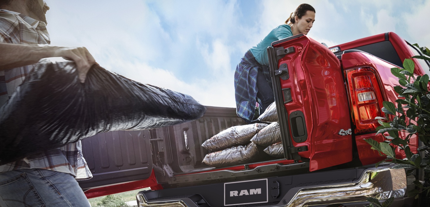 La caja de la camioneta en una Ram 1500 Tungsten 4x4 Crew Cab 2026 con la puerta trasera con división abierta y una mujer cargando bolsas de material en su caja.