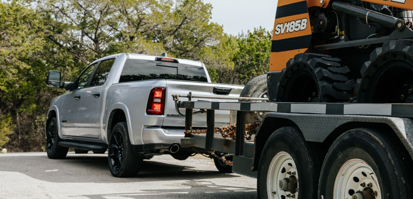 Un ángulo trasero de una Ram 1500 Laramie 4x4 Crew Cab 2026 blanca acarreando equipos motorizados en un remolque utilitario.